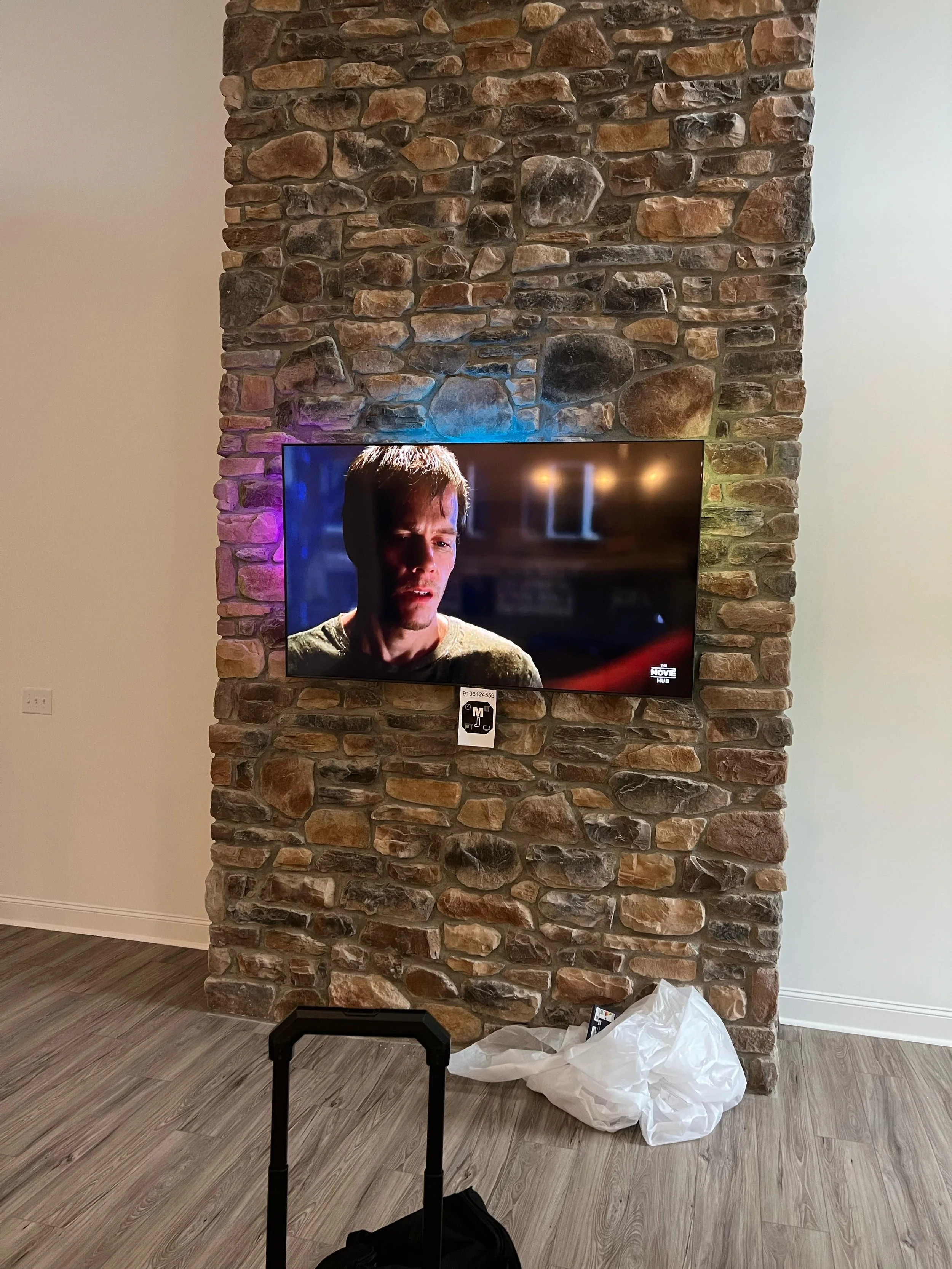 A stone fireplace with a mounted flat-screen TV displaying a scene from a movie. There is a black luggage handle in the foreground, a white plastic bag on the floor, and a small black object next to the bag.