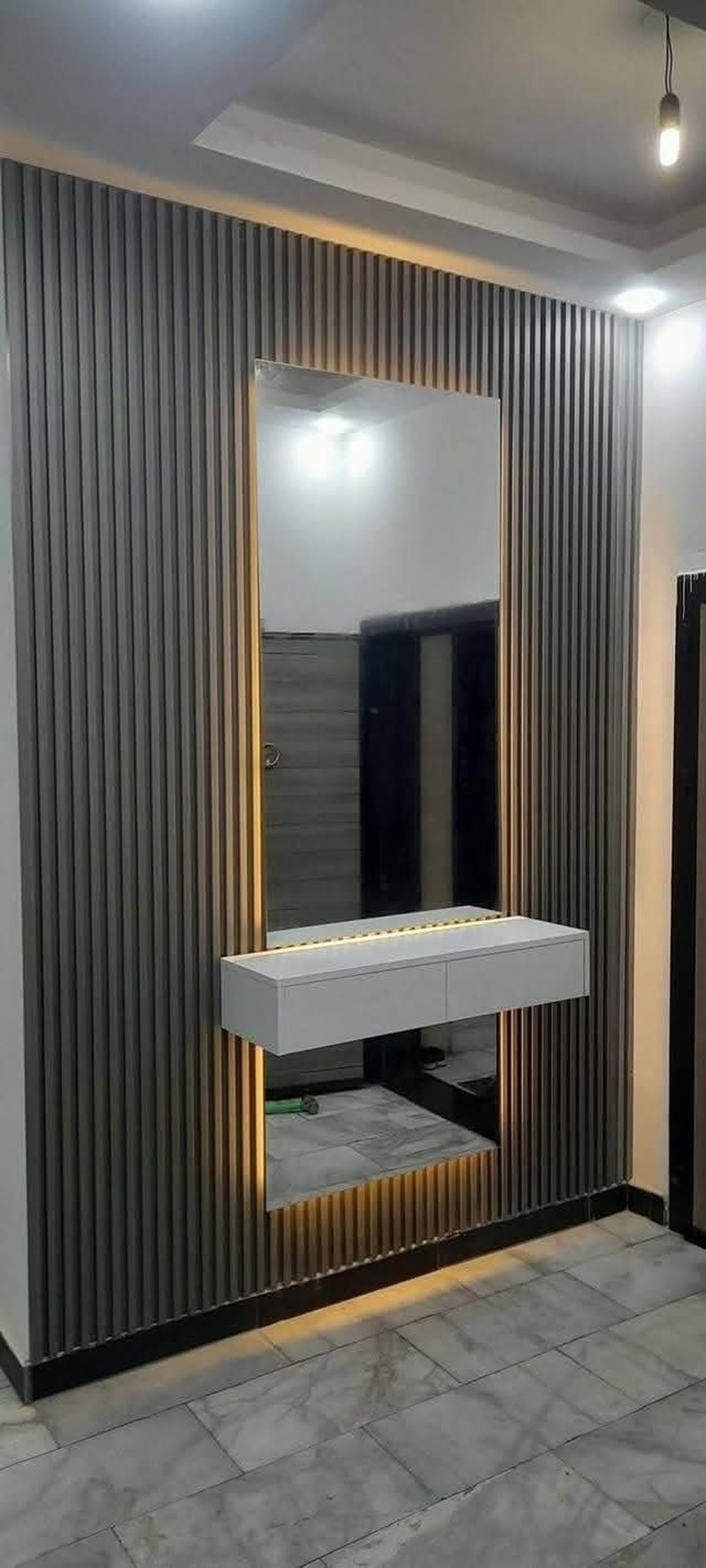 Modern hallway with a vertical ribbed wall, a large mirror, a white floating shelf, and gray tiled flooring.