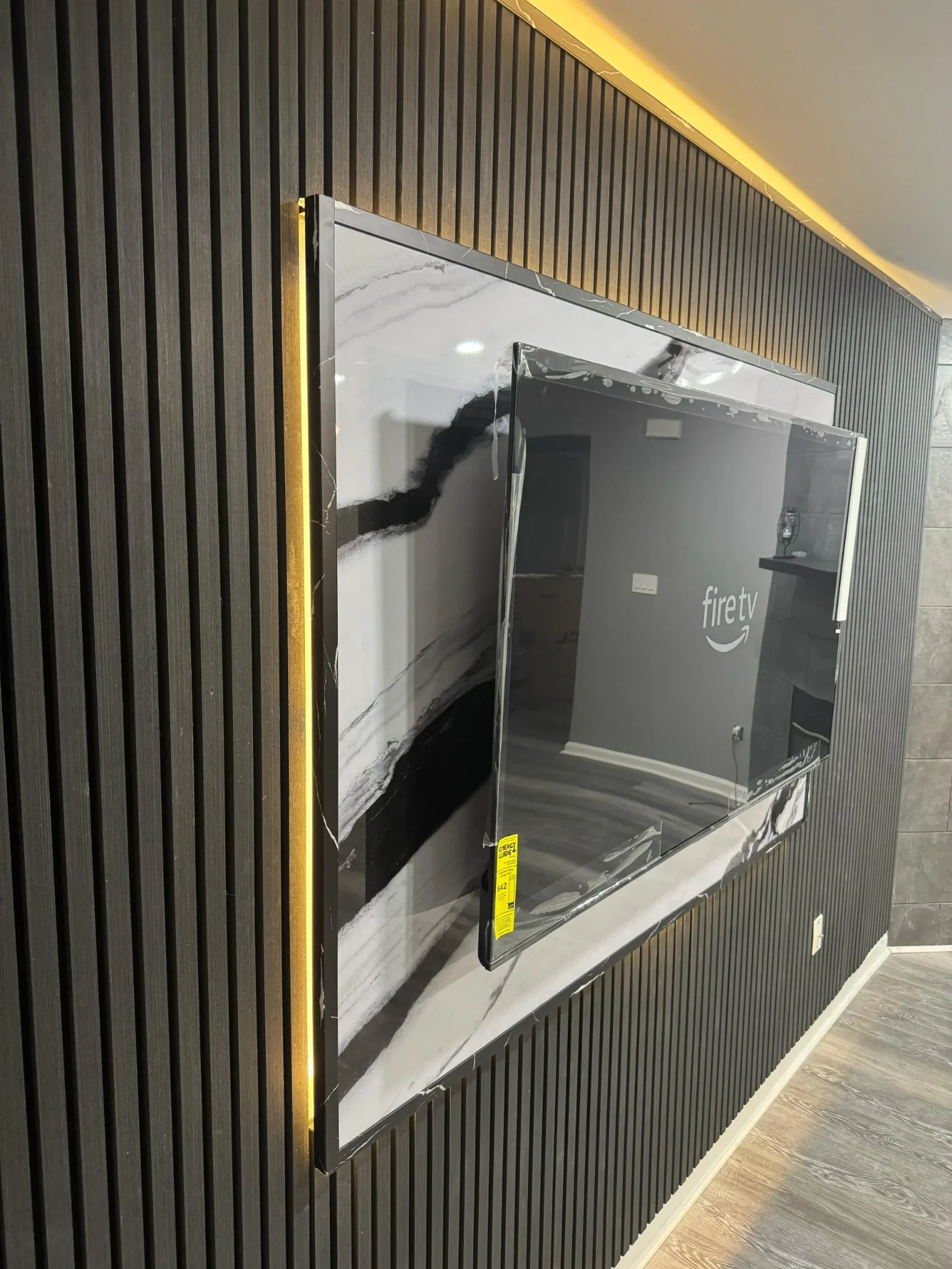A modern wall-mounted flat-screen TV in a room with black vertical slat wall paneling and wood flooring, reflecting a room with gray walls, a black fireplace, and a shelf with decorative items.