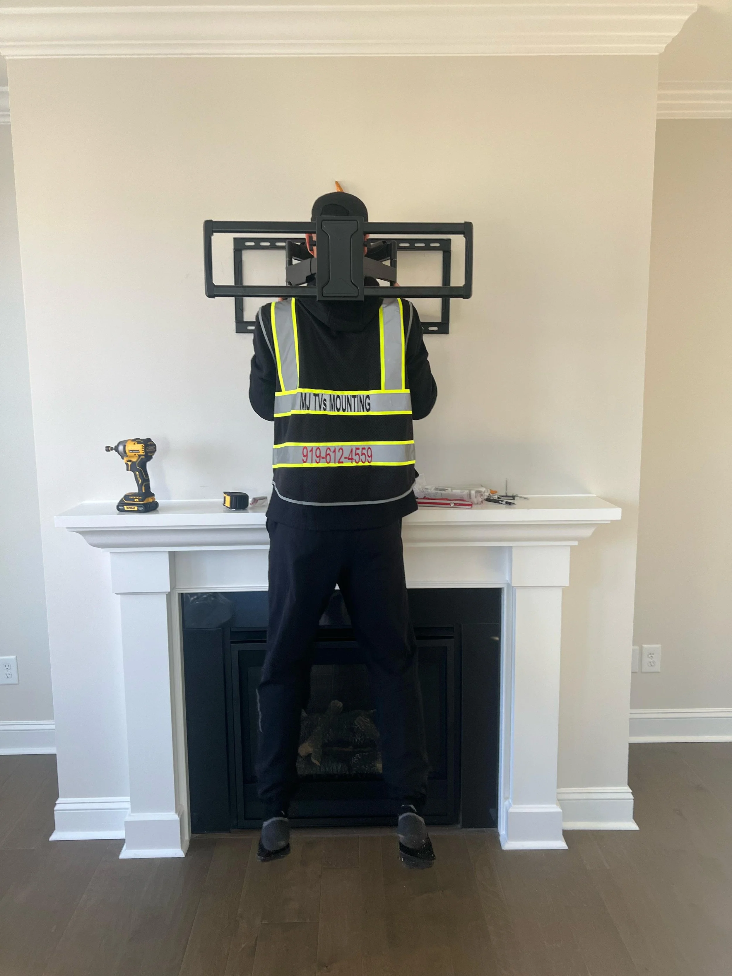 A person installing a wall-mounted TV above a fireplace, wearing a high-visibility vest, standing in front of a white wall in a living room.