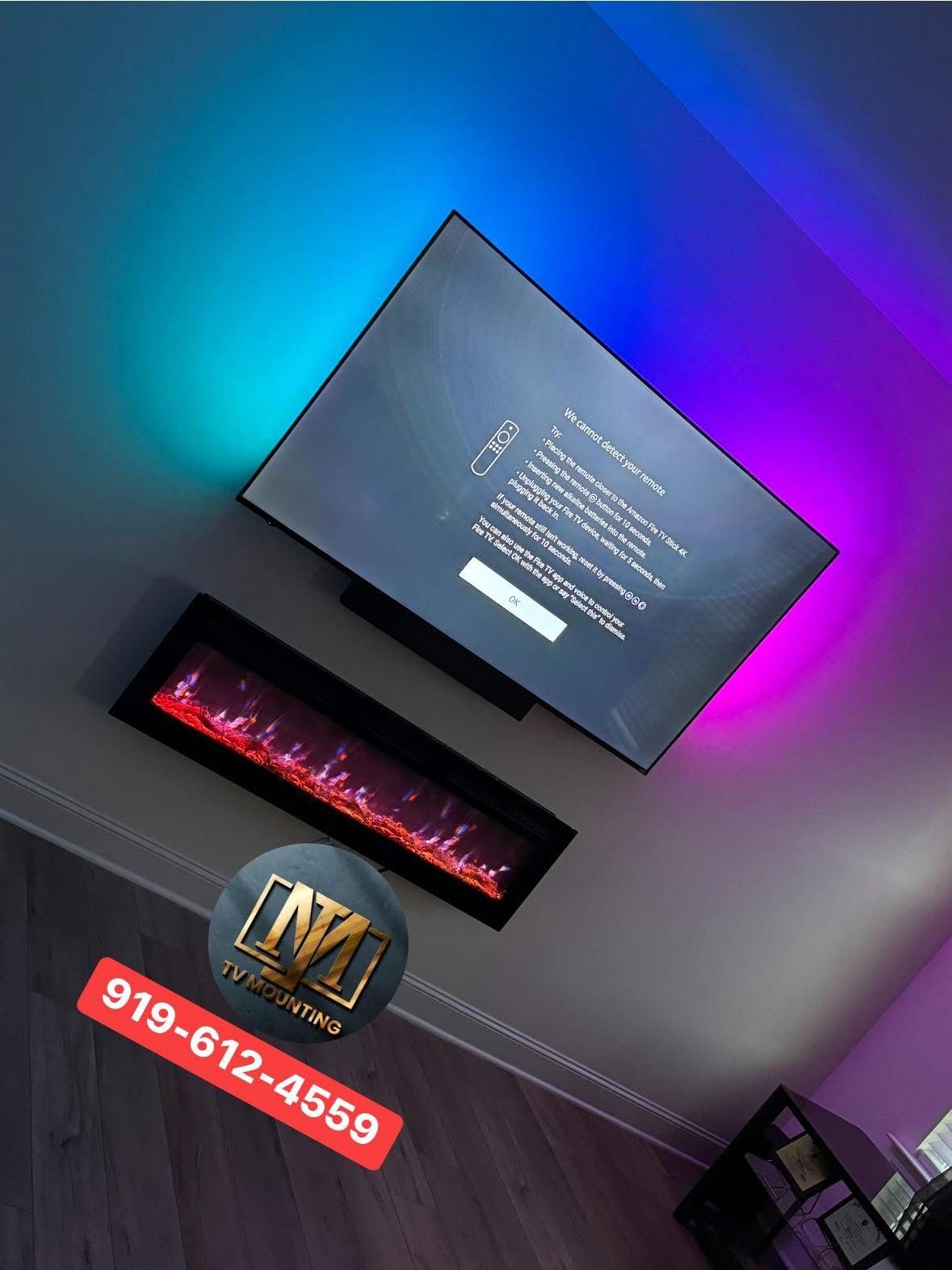 Living room with a mounted flat-screen TV displaying a message about remote detection issues, a modern electric fireplace underneath the TV, and a TV mounting service advertisement on the floor with a logo and phone number. Wall has colored ambient lighting.