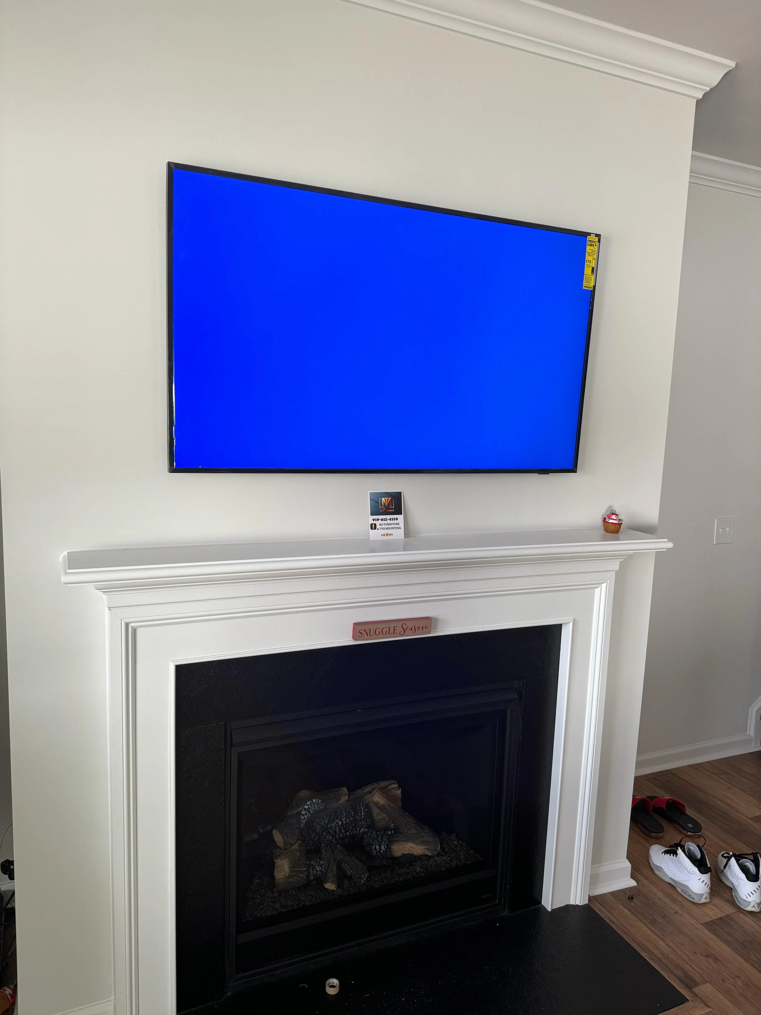 A living room with a white fireplace and a flat-screen television mounted above it. The TV screen is blue. There are small decorations on the fireplace mantel, including a card, a cupcake ornament, and a pink sign that reads 'SNUGGLE Season.' On the floor next to the fireplace, there are a pair of white sneakers and other shoes.