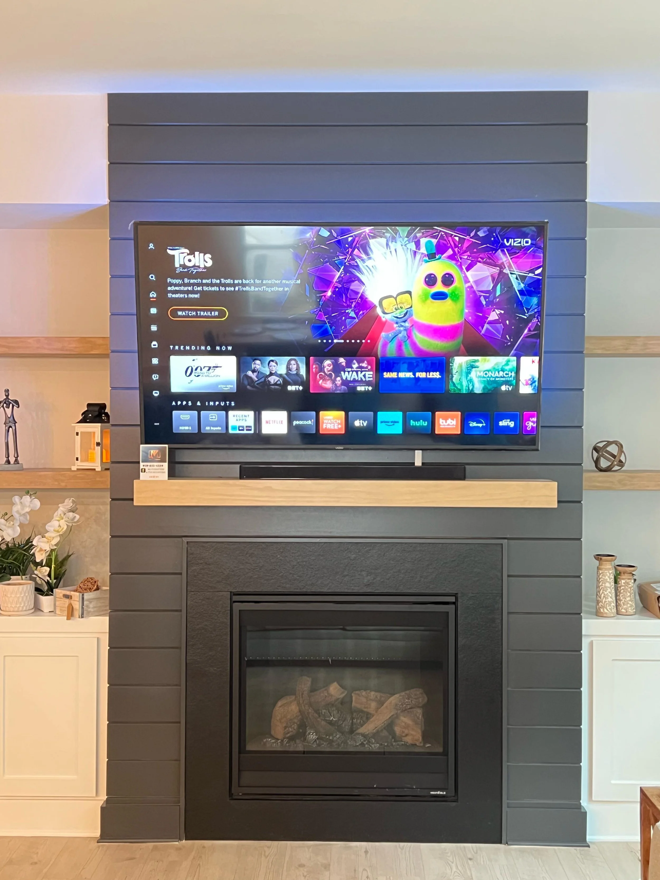 Living room fireplace with a mounted flat-screen TV above, displaying a streaming service menu. The fireplace has a glass front with decorative logs inside, surrounded by dark gray paneling. On either side of the fireplace, there are white side tables with decorative items and plants.