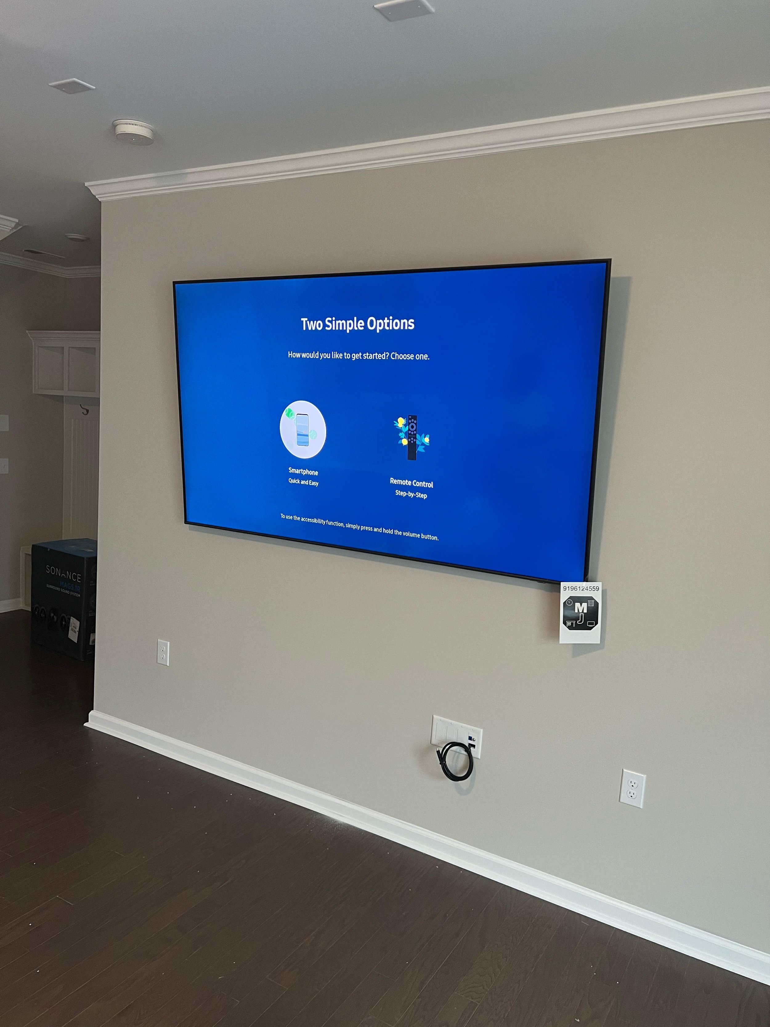 Wall-mounted TV screen displaying set-up options, with a power outlet and wall connections below, in a modern room with hardwood flooring.
