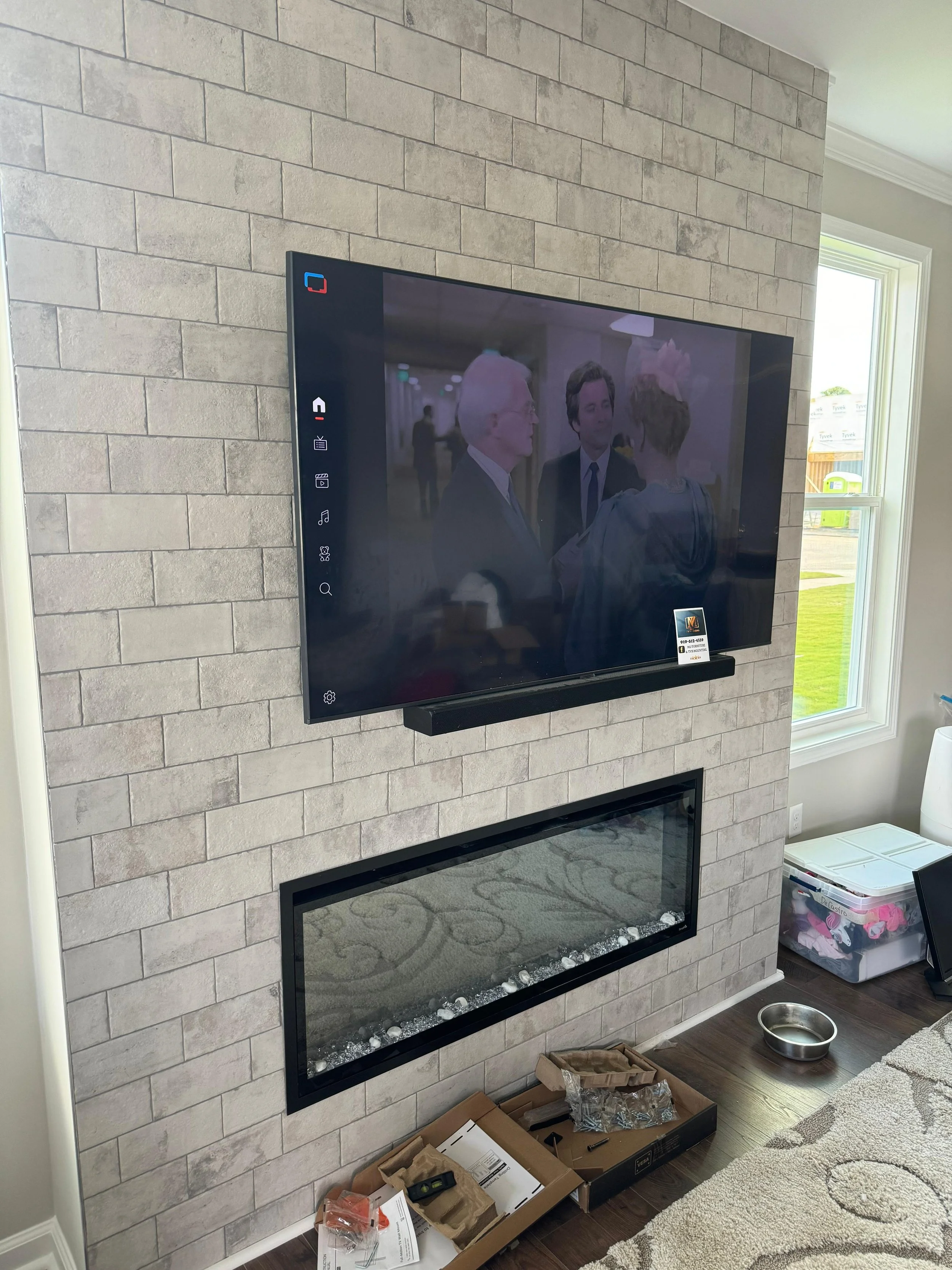 Living room fireplace with a wall-mounted flat-screen TV above it, brick wall behind, next to a window with a view of a yard. Items on the floor include boxes and a dog bowl.