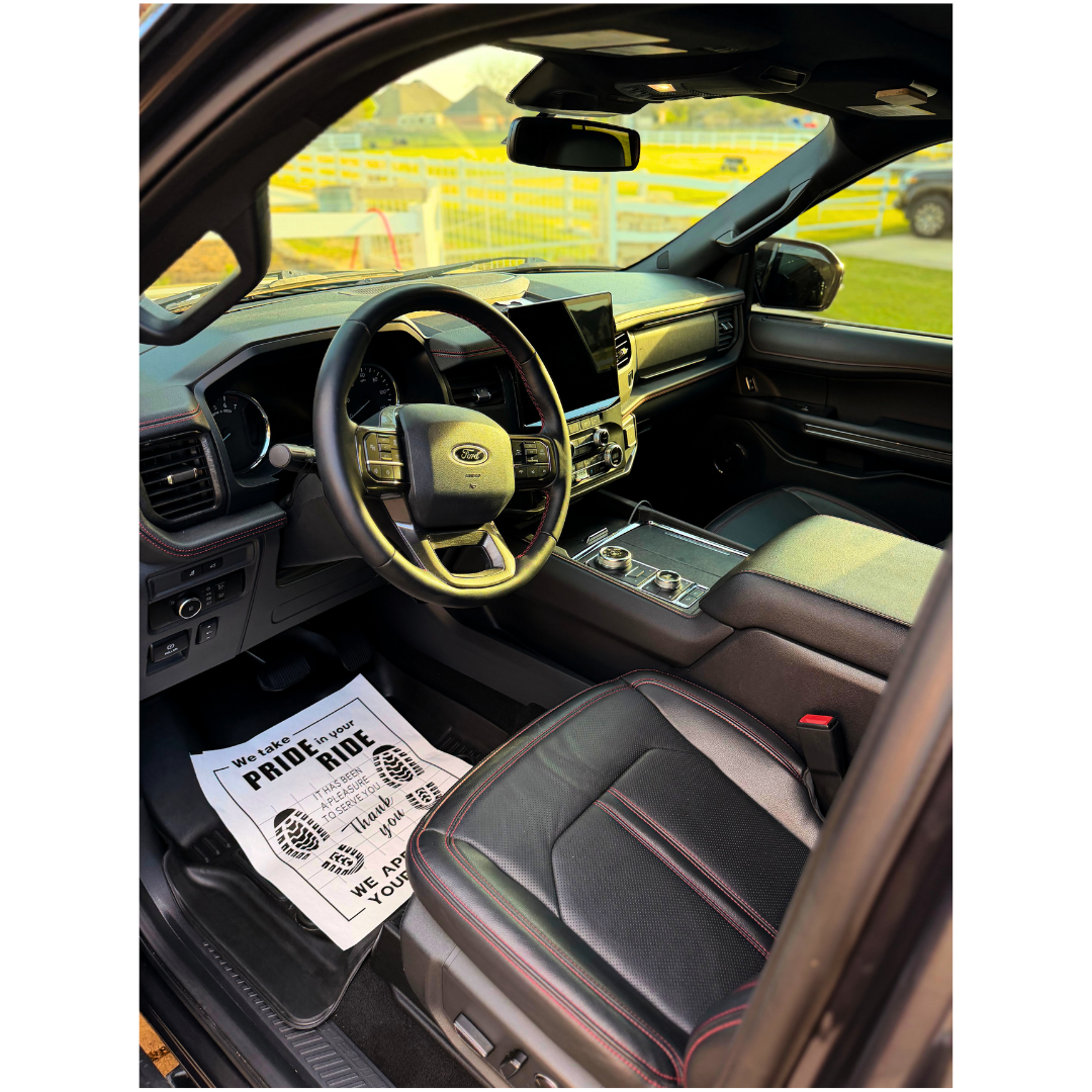 Interior of a Ford vehicle with a black leather seat with red stitching, steering wheel, dashboard, digital display, and a paper floor mat with text about pride in riding and gratitude.