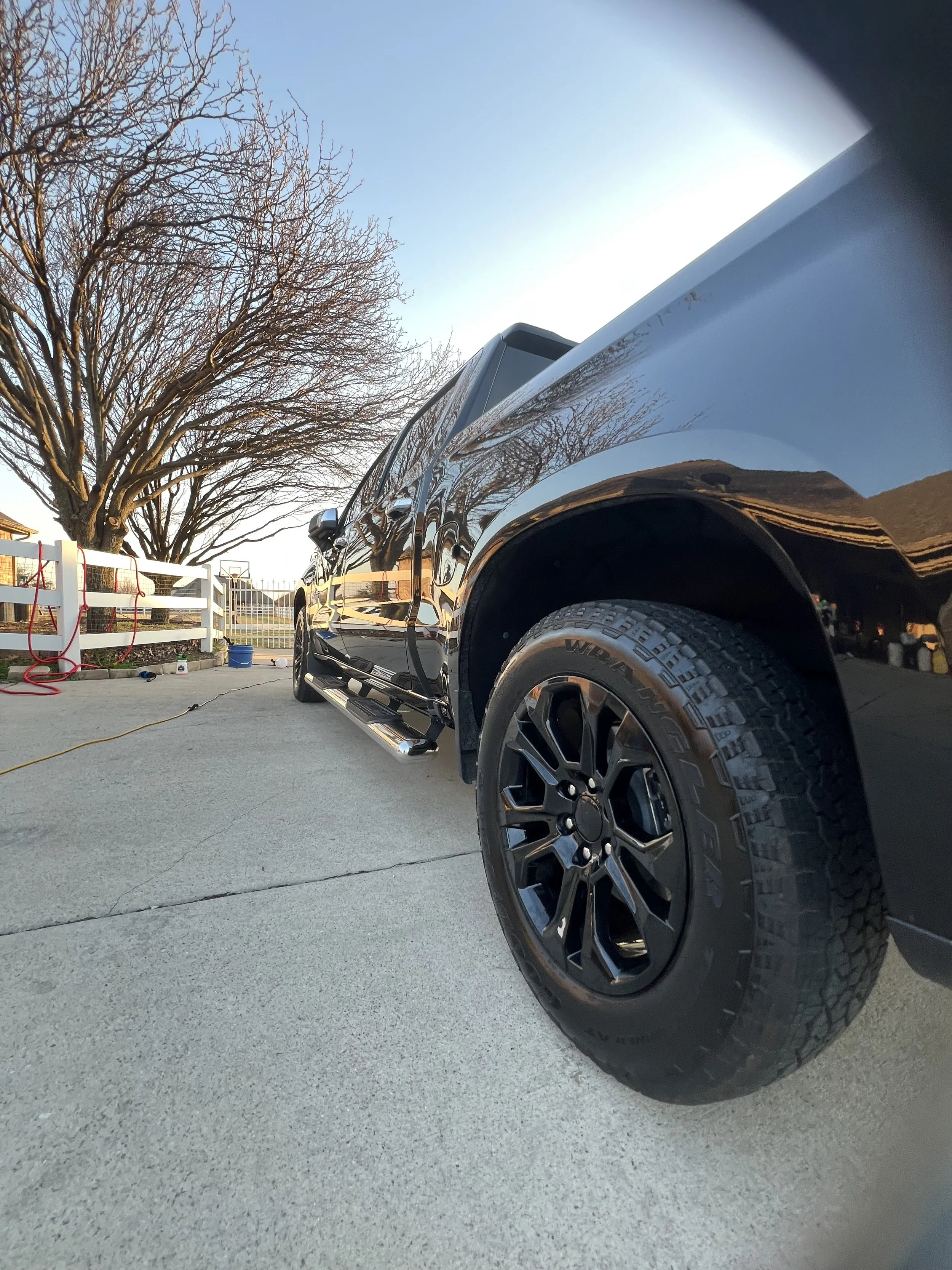 A black pickup truck parked on a driveway near a tree with bare branches and a white fence. The truck is shiny and reflects the surroundings.