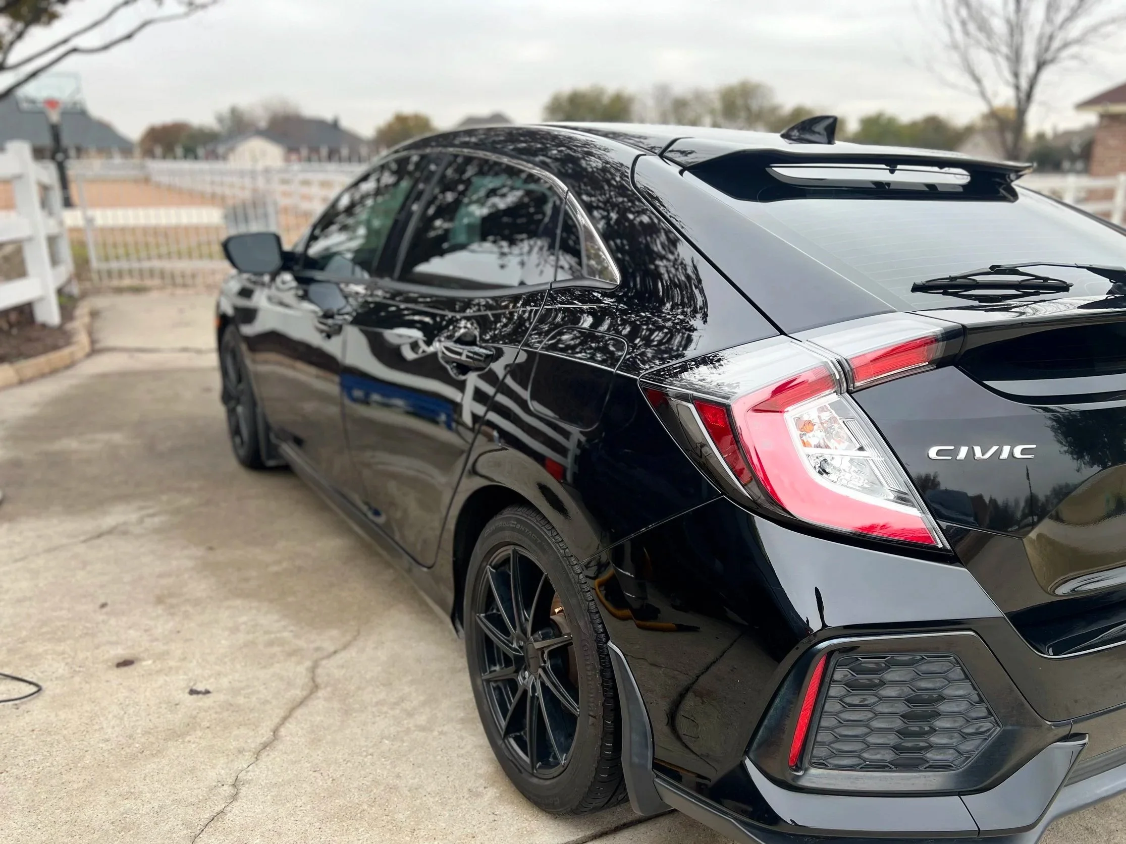A black Honda Civic parked on a driveway with a white fence, houses, trees, and cloudy sky in the background.