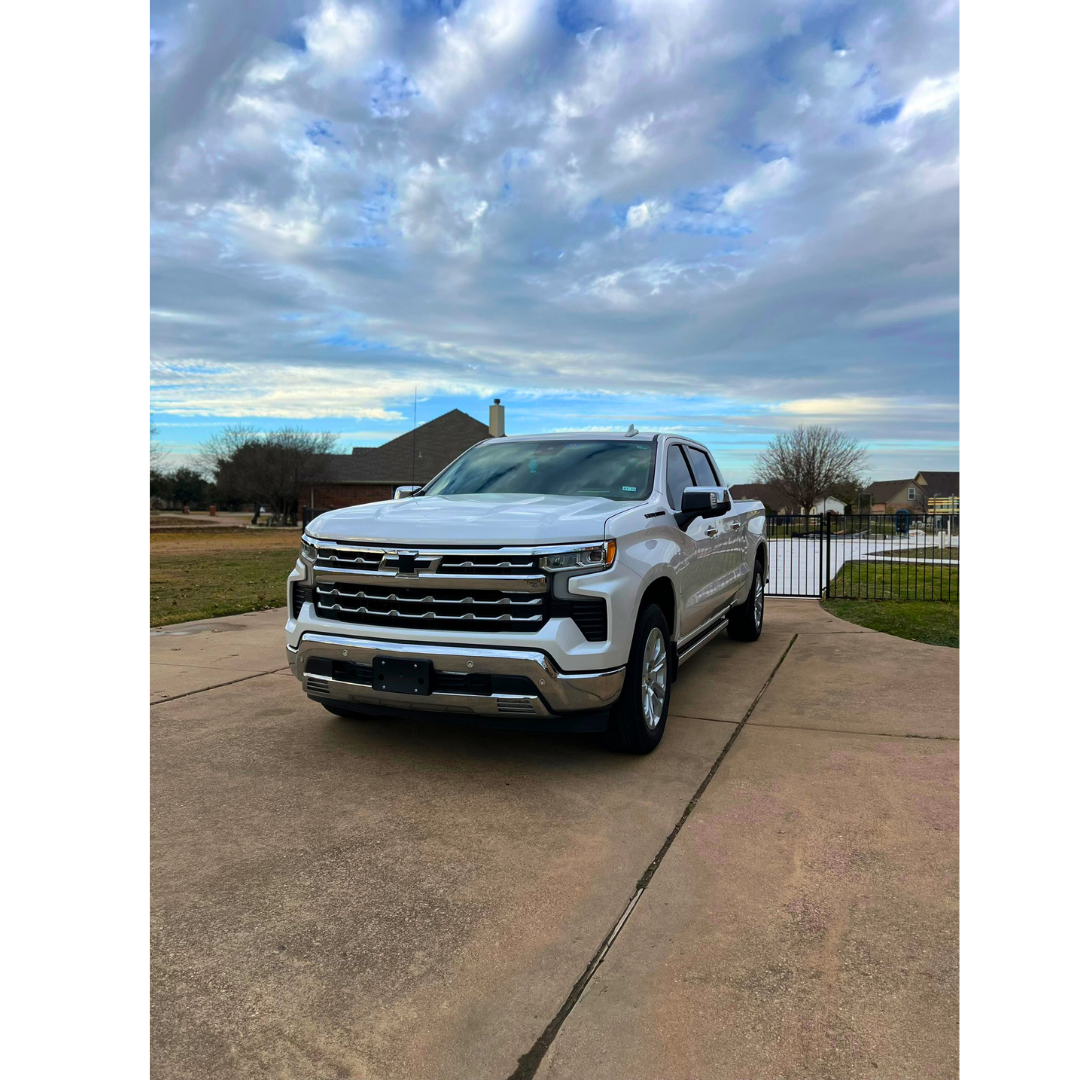 A white Chevrolet pickup truck parked on a concrete driveway in a suburban neighborhood with a cloudy sky overhead.