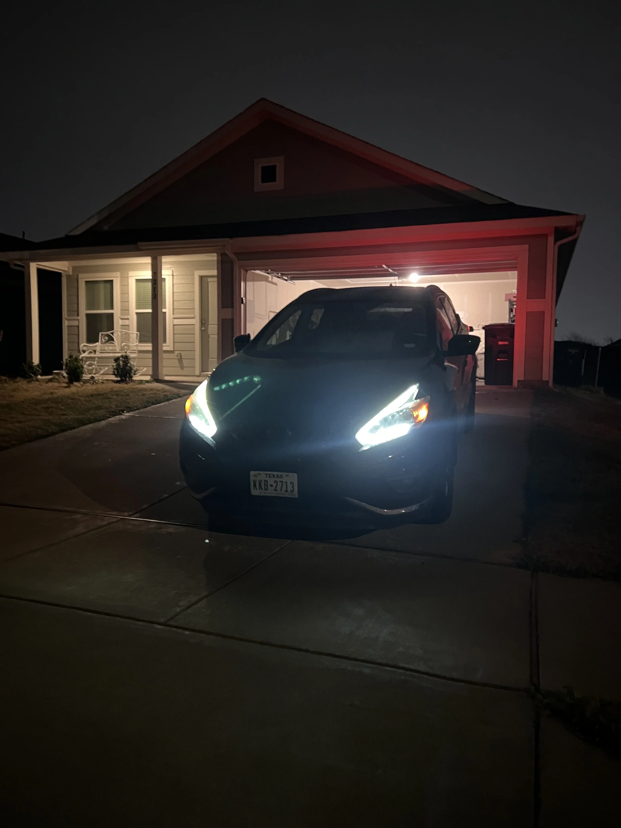 A black car with bright headlights parked in front of a house garage at night, with the interior lights of the garage turned on.