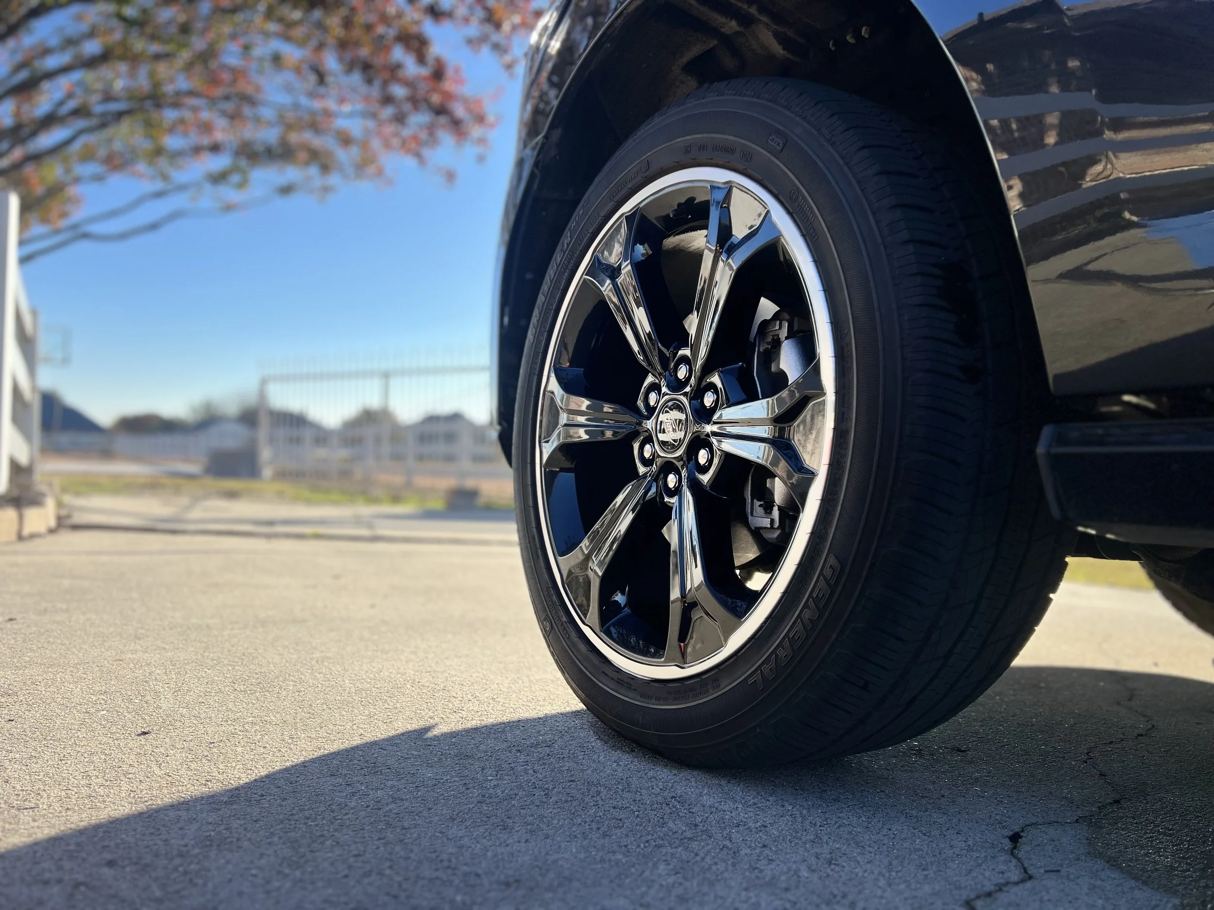 Close-up of a car wheel with a shiny black and chrome rim and a Goodyear Generals tire, on a concrete surface outdoors with a fence, trees, and houses in the background.