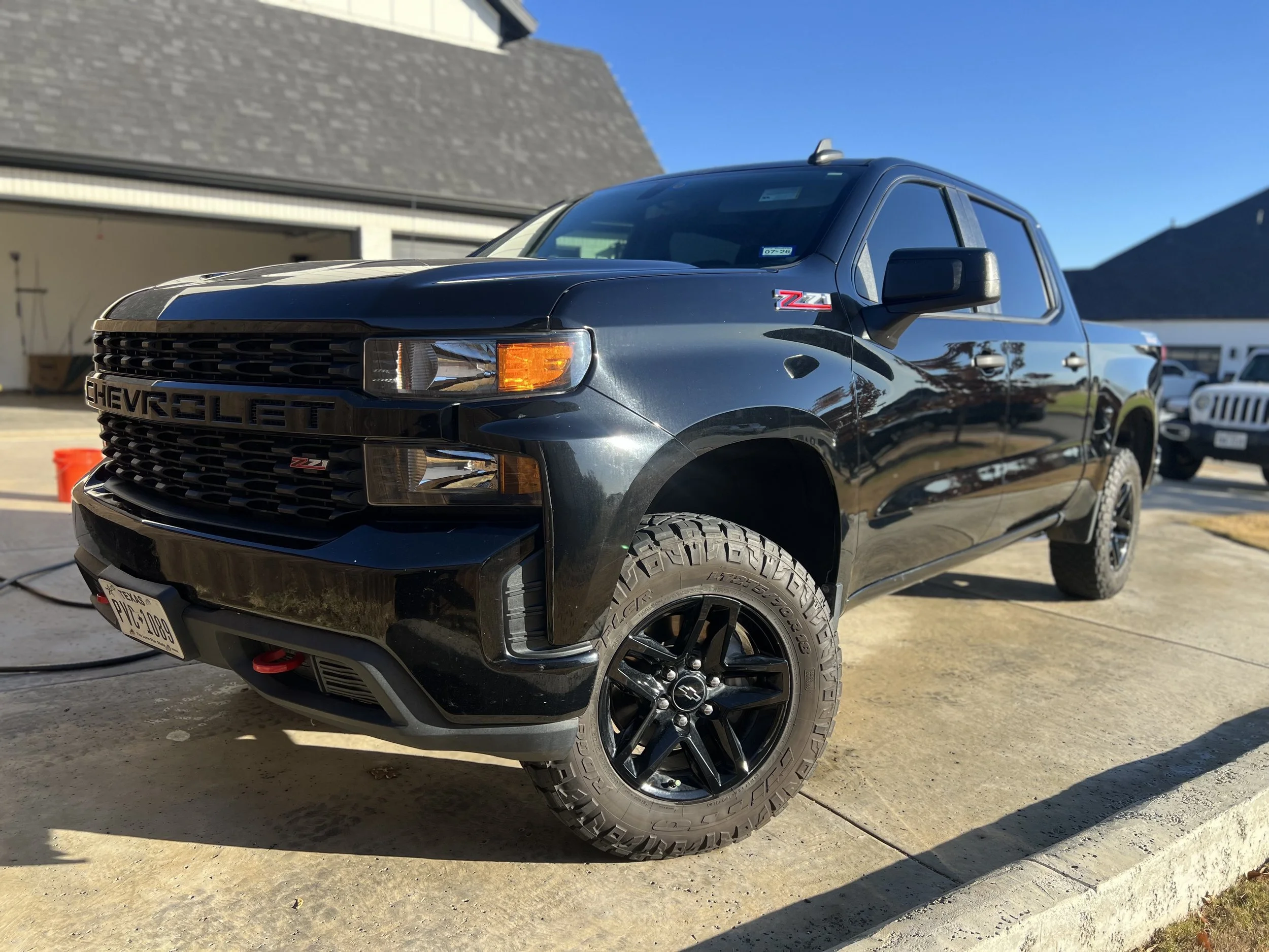 Black Chevrolet pickup truck parked on driveway with a garage and other vehicles in the background, under a clear blue sky.