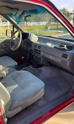 Interior of a car showing the driver's seat, steering wheel, dashboard, and front passenger seat, with the window rolled down and scenery outside.
