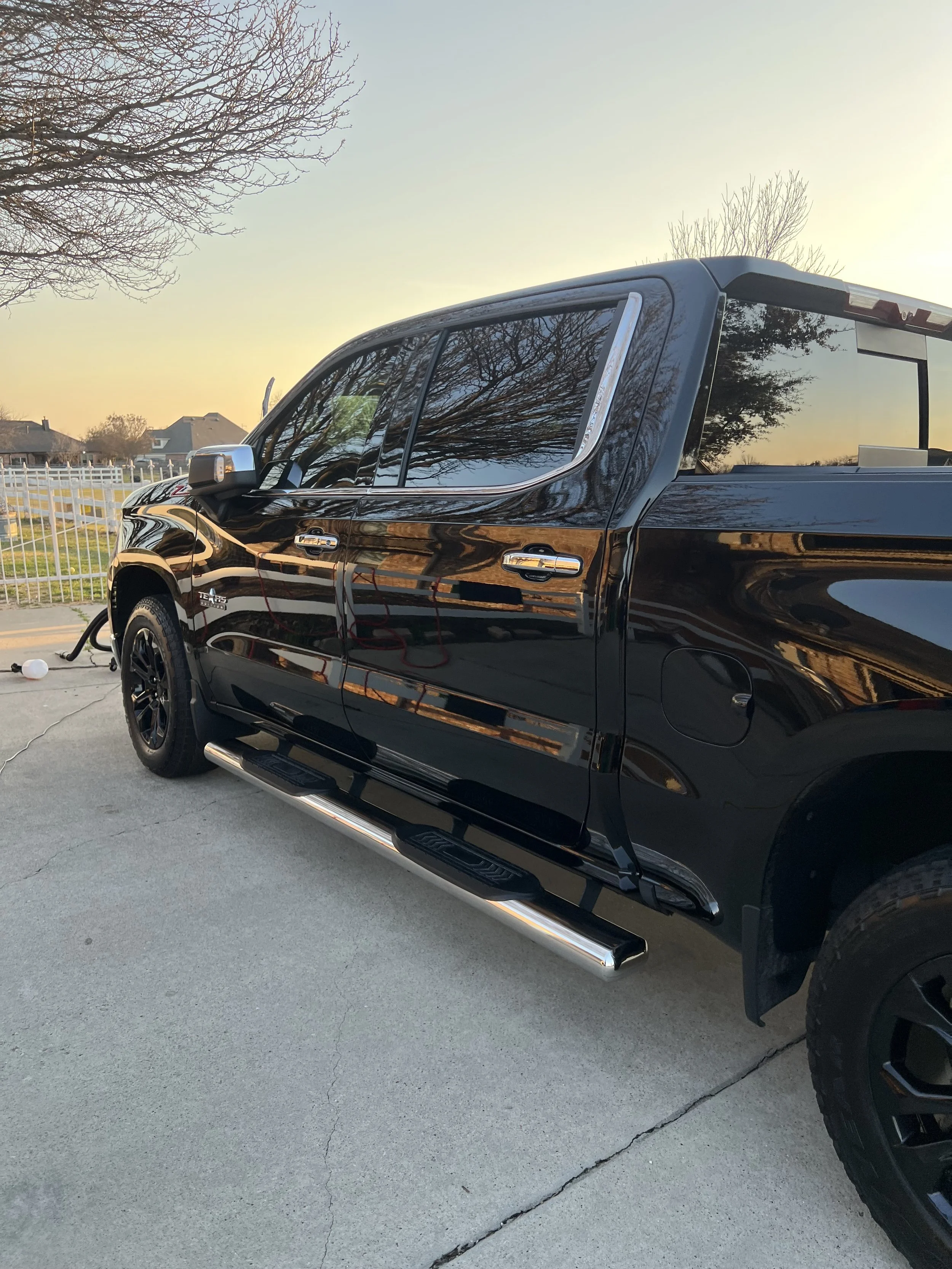 A black pickup truck parked outside on a concrete surface during sunset, with reflections of trees and sky on its surface.