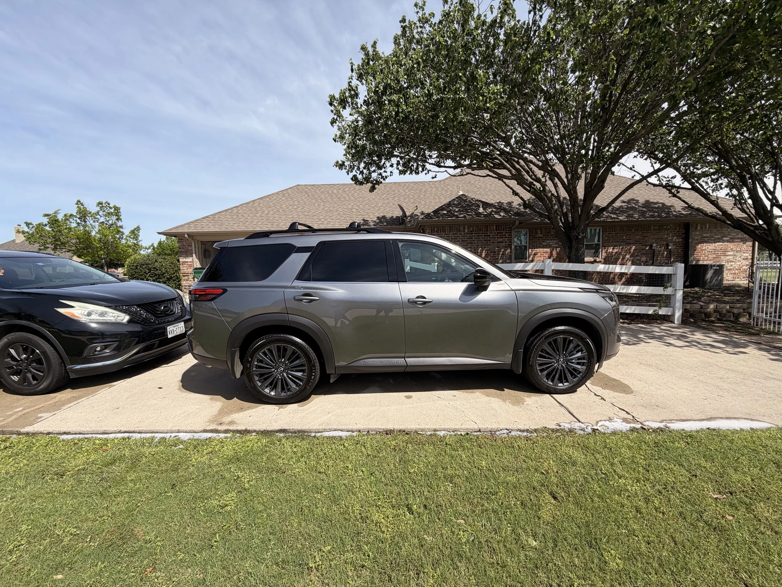 A silver SUV parked on a driveway next to a black sedan in front of a brick house with trees and a white fence.