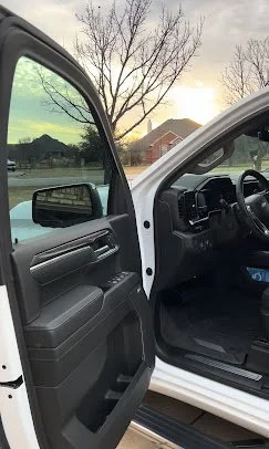 Open car door showing the interior dashboard and side panel, with a tree and houses outside in the background during sunset.
