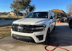 A white SUV parked on a residential street with trees and houses in the background.