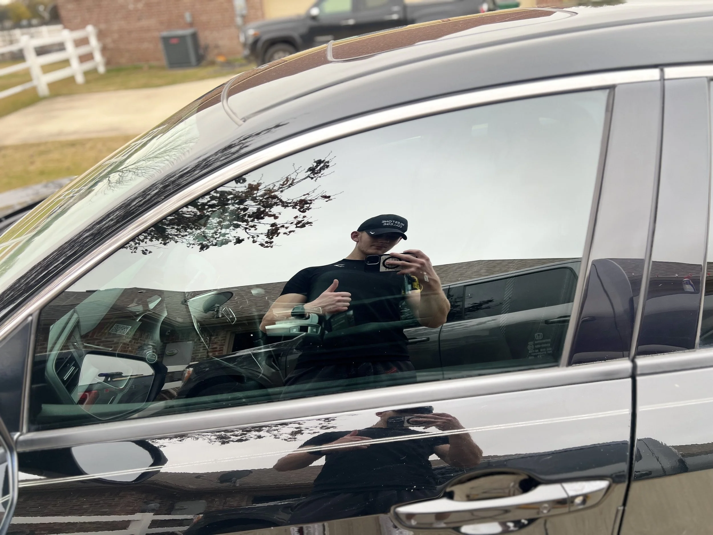Reflection of a person taking a photo in the car's window, with the person's upper body, black shirt, and black cap visible.