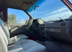 Inside a vehicle showing the driver's seat, steering wheel, dashboard, and windshield with trees and blue sky outside.