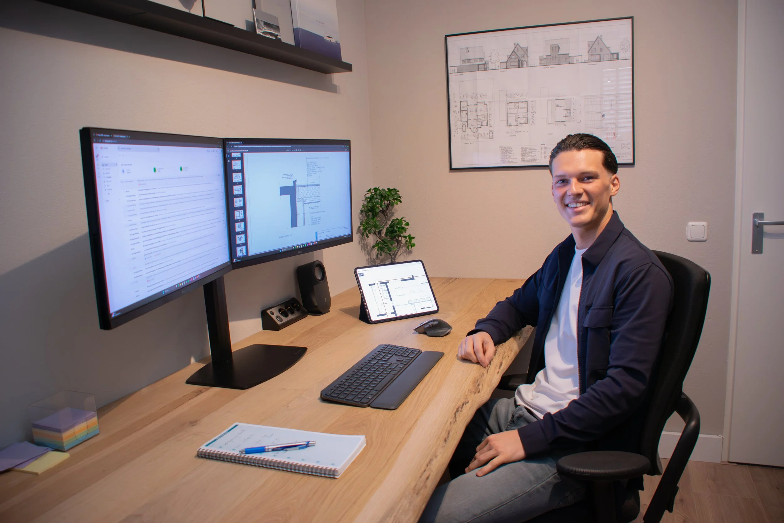 A man sitting at a wooden desk with dual monitors, a tablet, and a keyboard, smiling at the camera inside a home office.