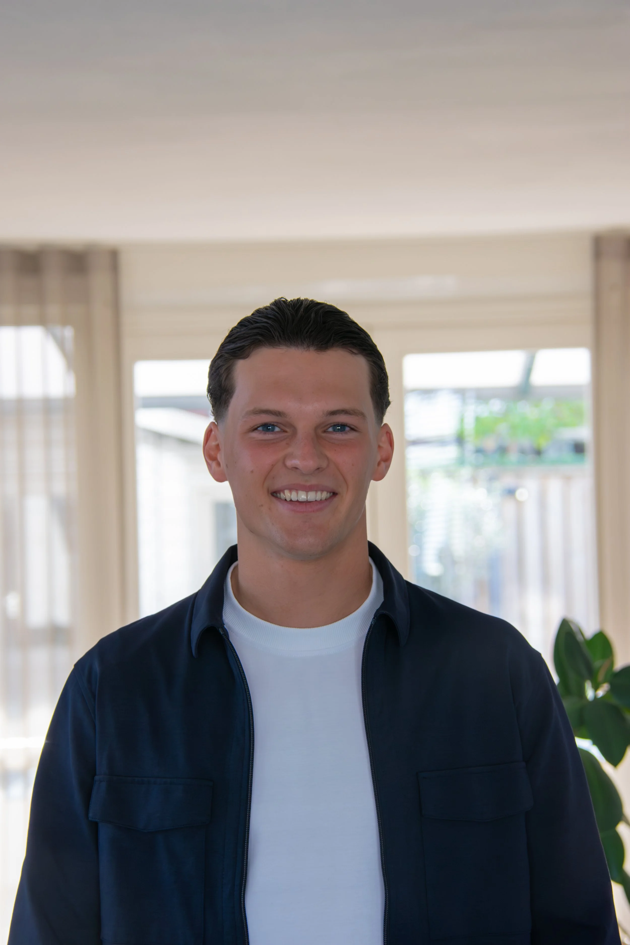 Young man with dark hair, wearing a white t-shirt and dark jacket, smiling indoors near large windows and a green plant.