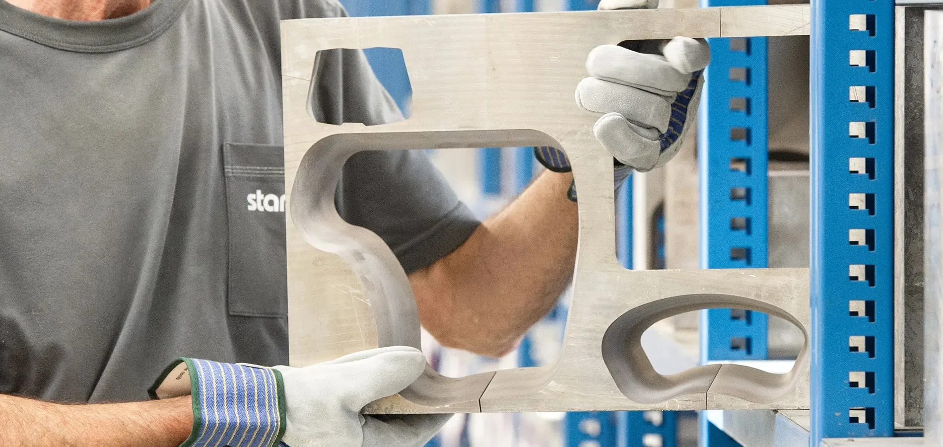 A person wearing a gray shirt with a pocket and a glove on their left hand is holding a rectangular, metallic part with a curved section and openings, possibly made of aluminum, in a workshop or manufacturing setting. The person is seen through a cut-out window in the part. Blue shelving units are visible in the background.