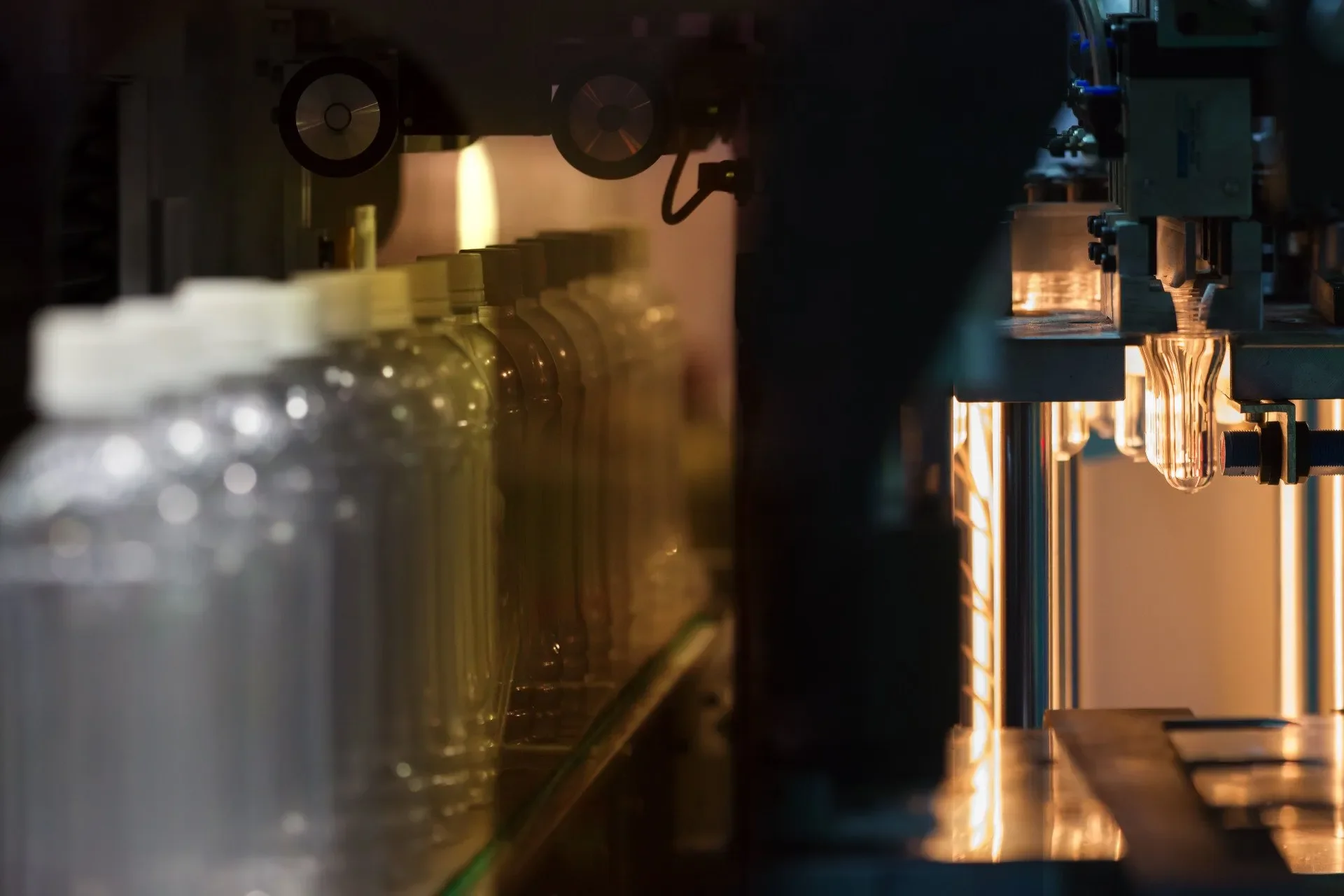 Close-up of a manufacturing or laboratory process involving glass containers and machinery, with glass bottles or test tubes aligned on the left and illuminated mechanical parts on the right.