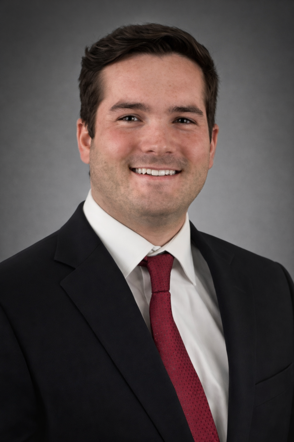 Headshot of a young man with dark hair, wearing a black suit, white shirt, and red tie, smiling against a gray background.