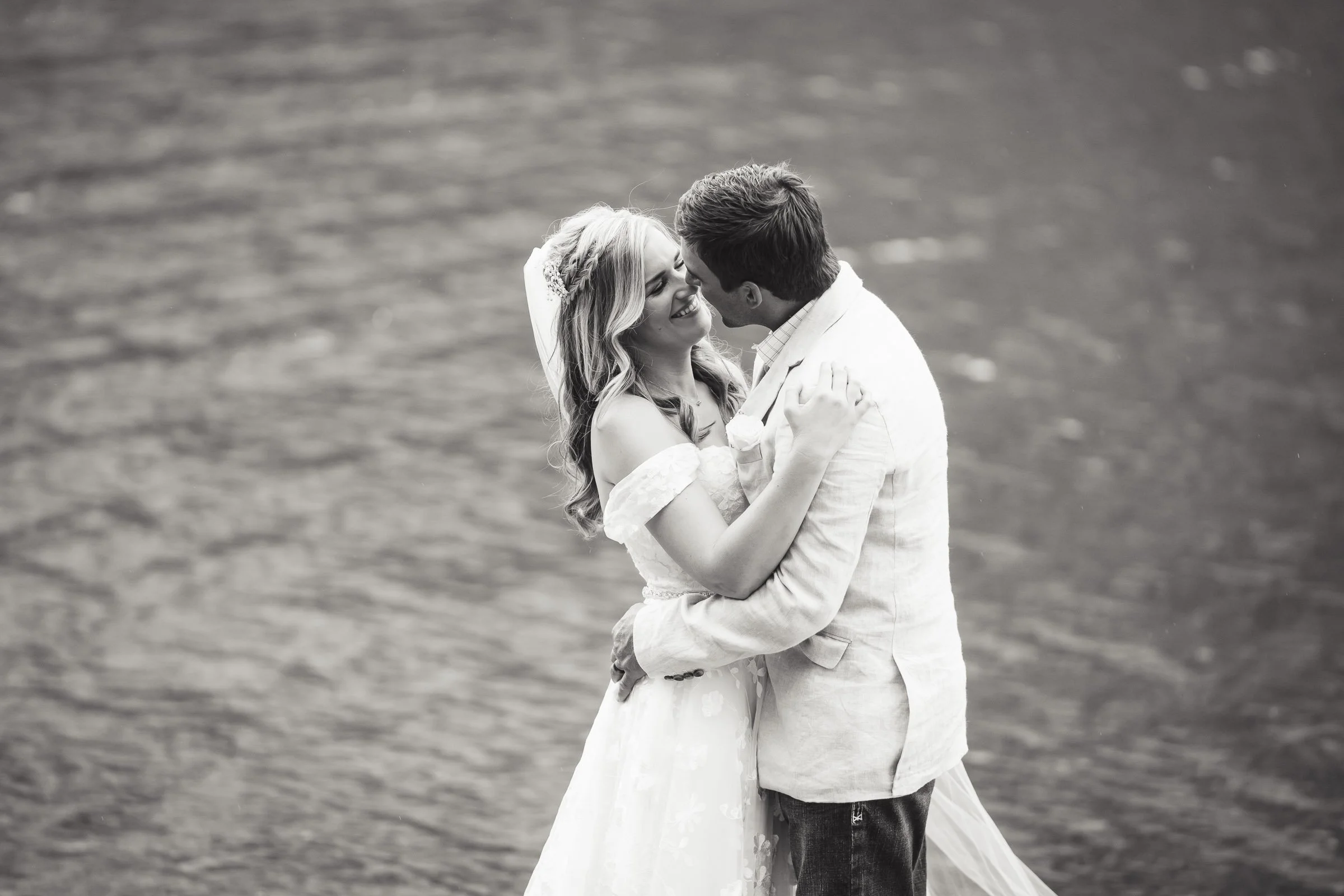 A black and white photo of a bride and groom hugging and about to kiss near a body of water.