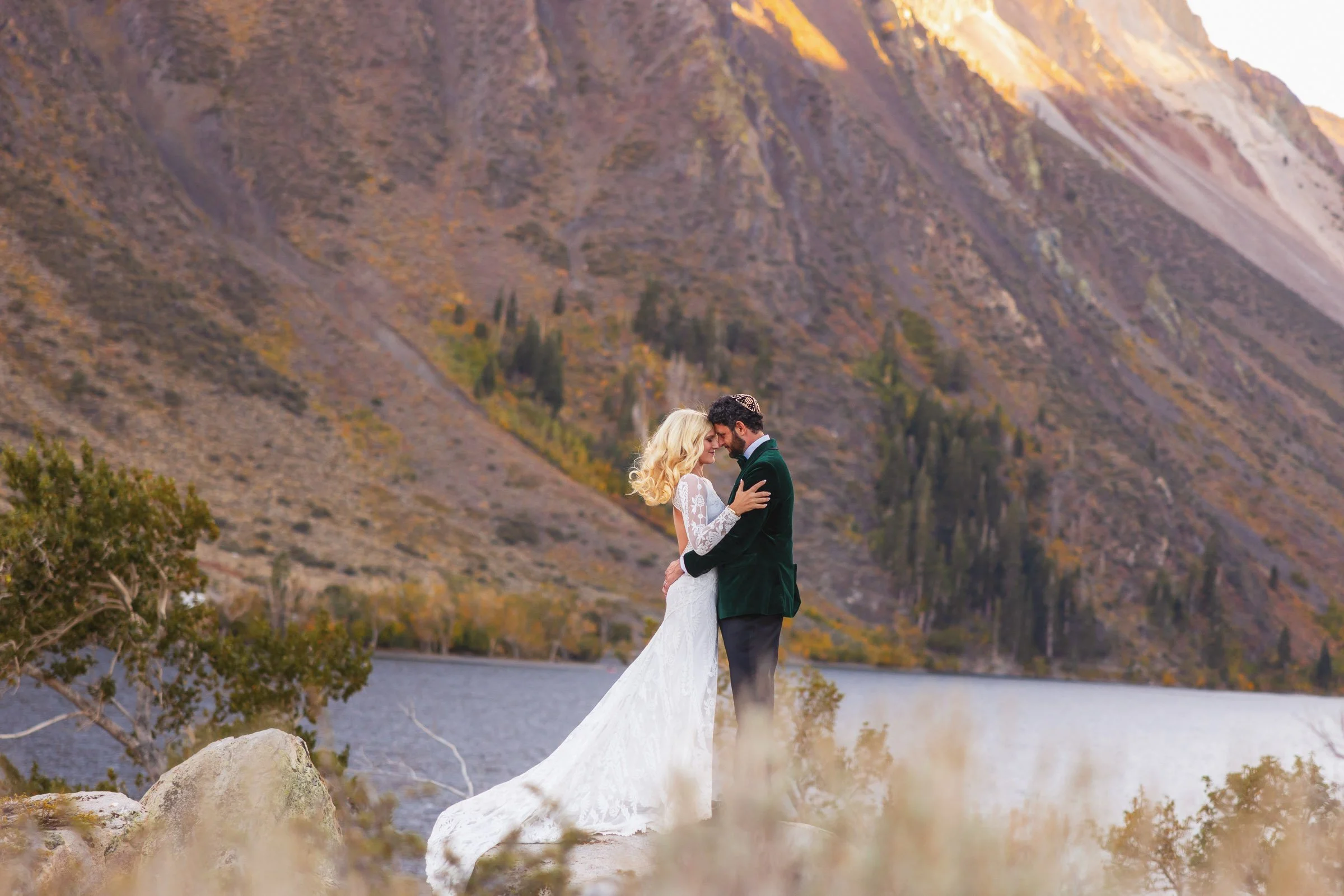 A bride and groom standing close together by a lake with mountains in the background.