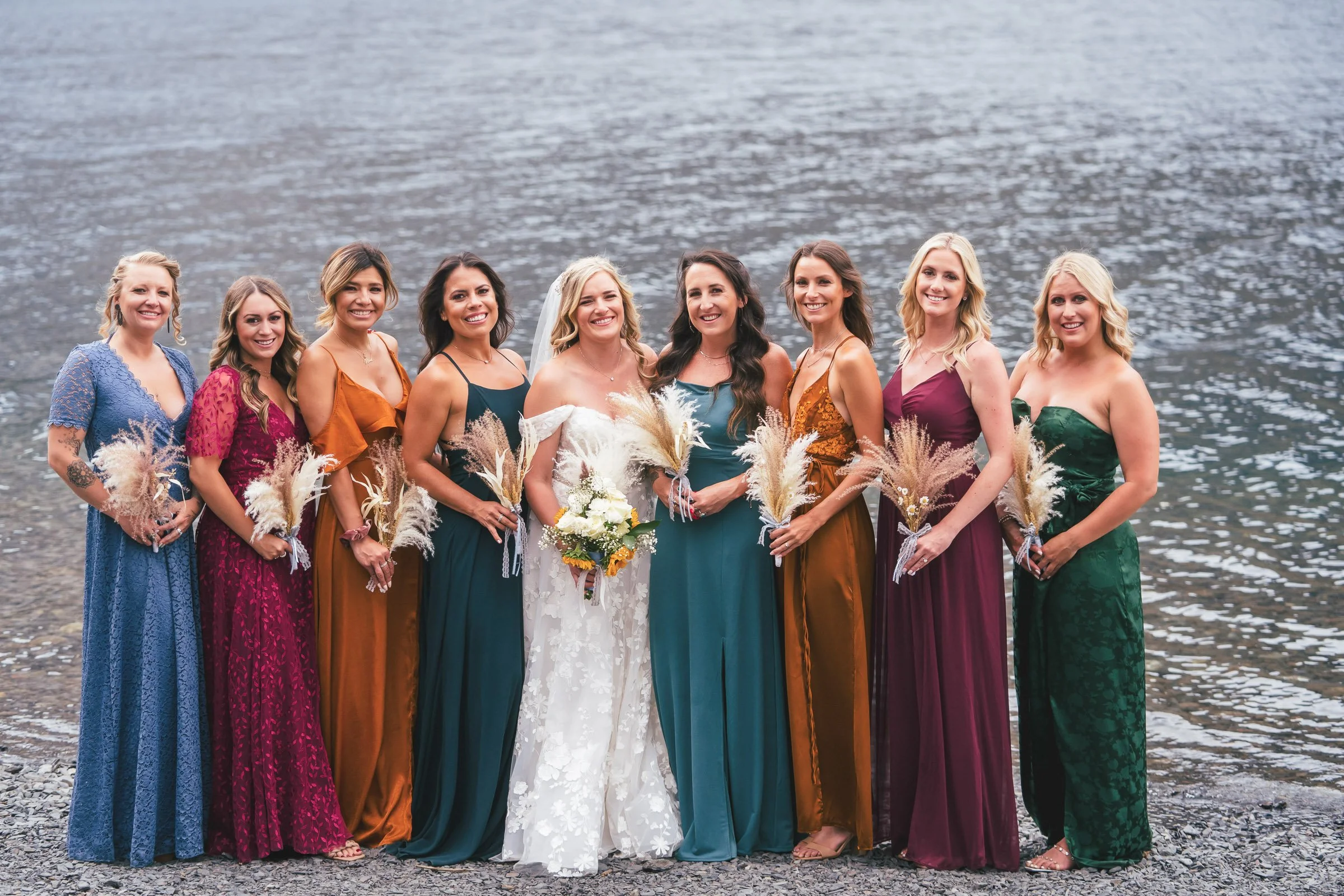 Women in colorful dresses standing by a body of water, celebrating a wedding with one woman in a white wedding gown and others in long jewel-toned dresses, holding bouquets.