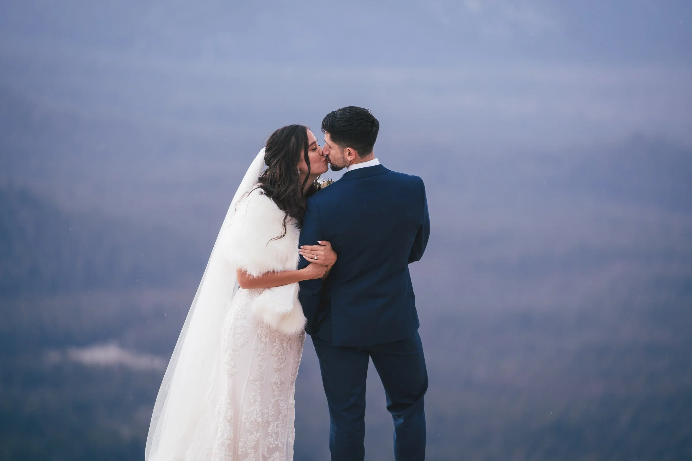 A bride and groom sharing a kiss outdoors against a cloudy sky, dressed in wedding attire.