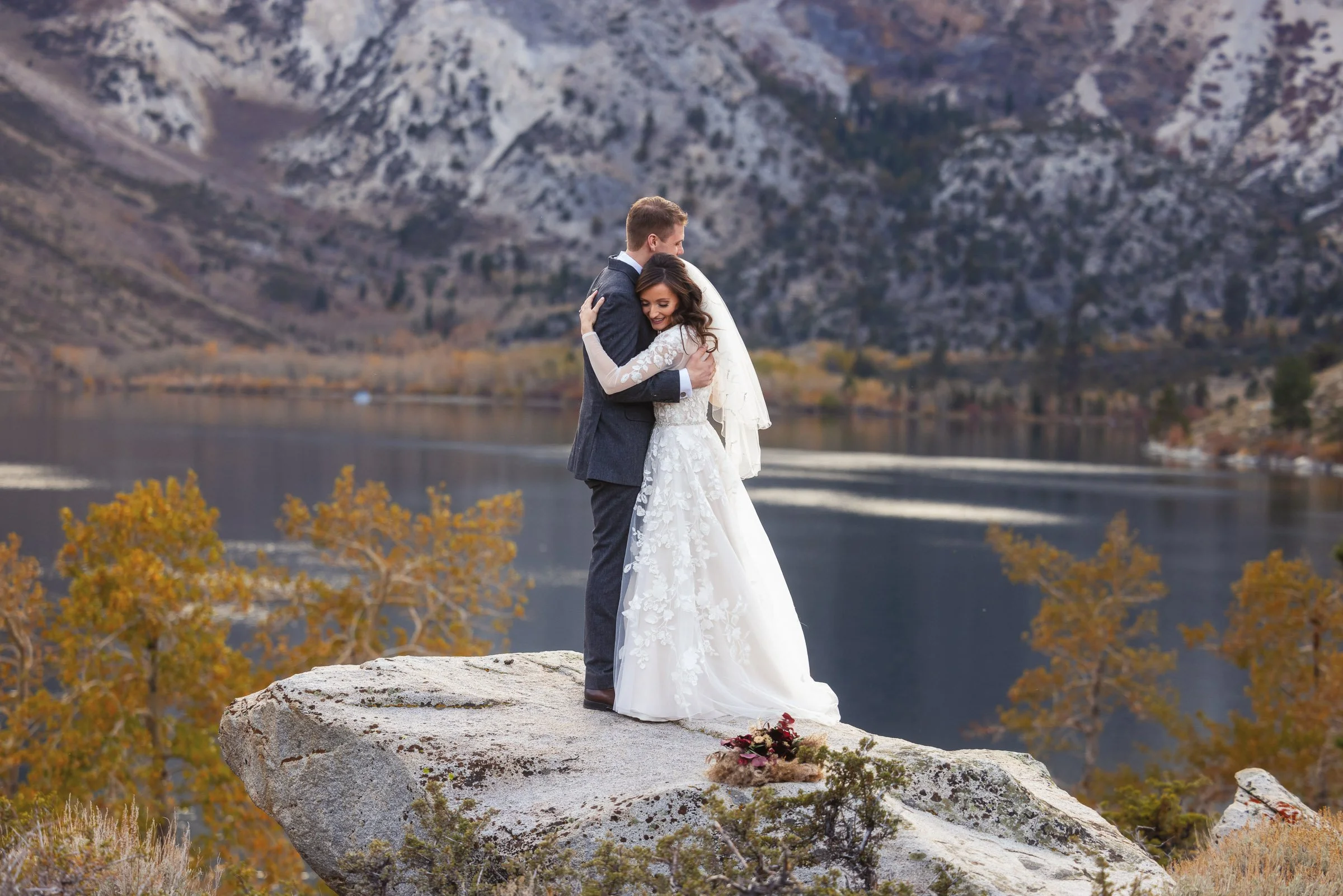 A newlywed couple hugging on a large rock by a lake with mountains and autumn trees in the background.