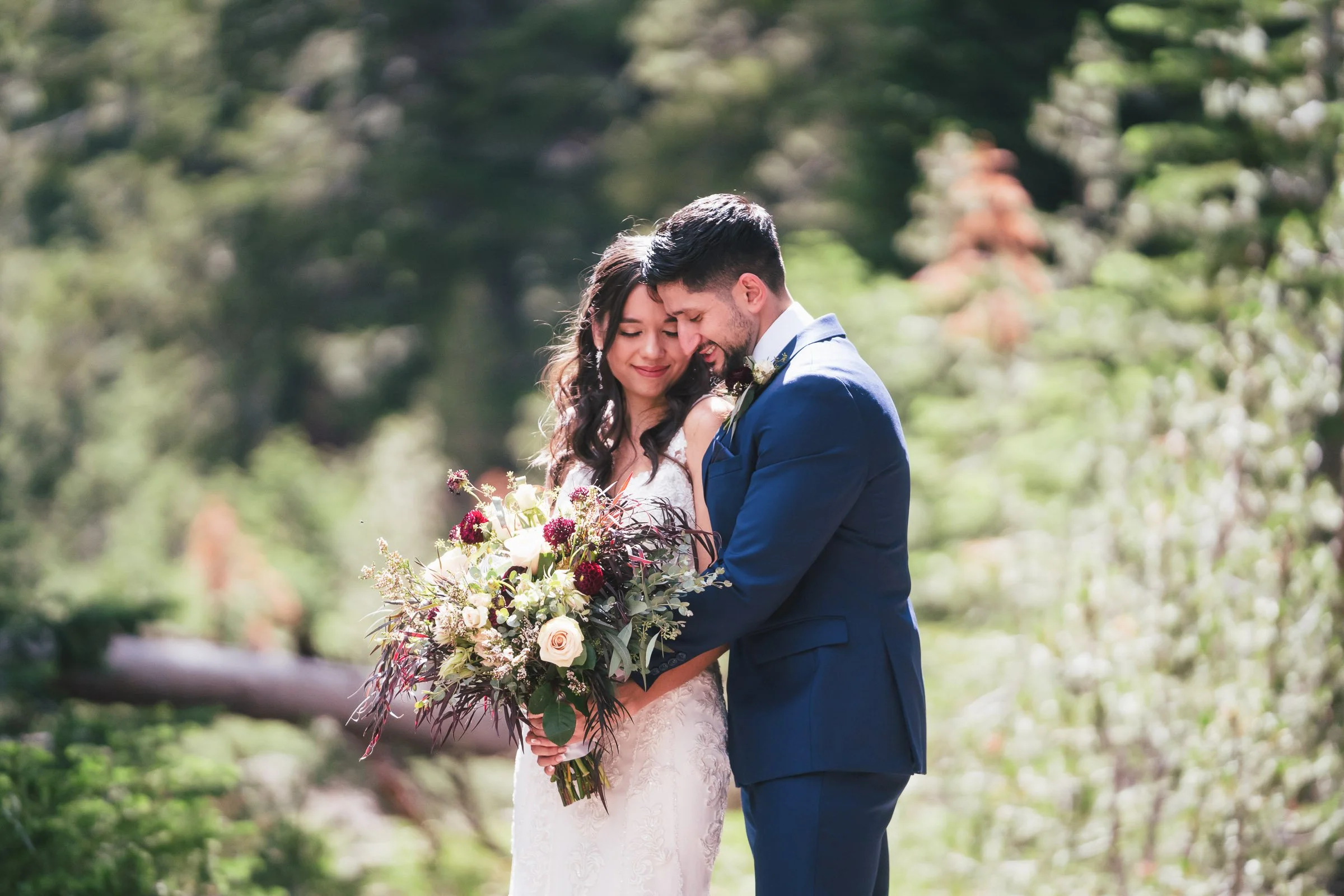 A bride and groom holding a bouquet of flowers, embracing each other with smiling faces in a lush outdoor setting.