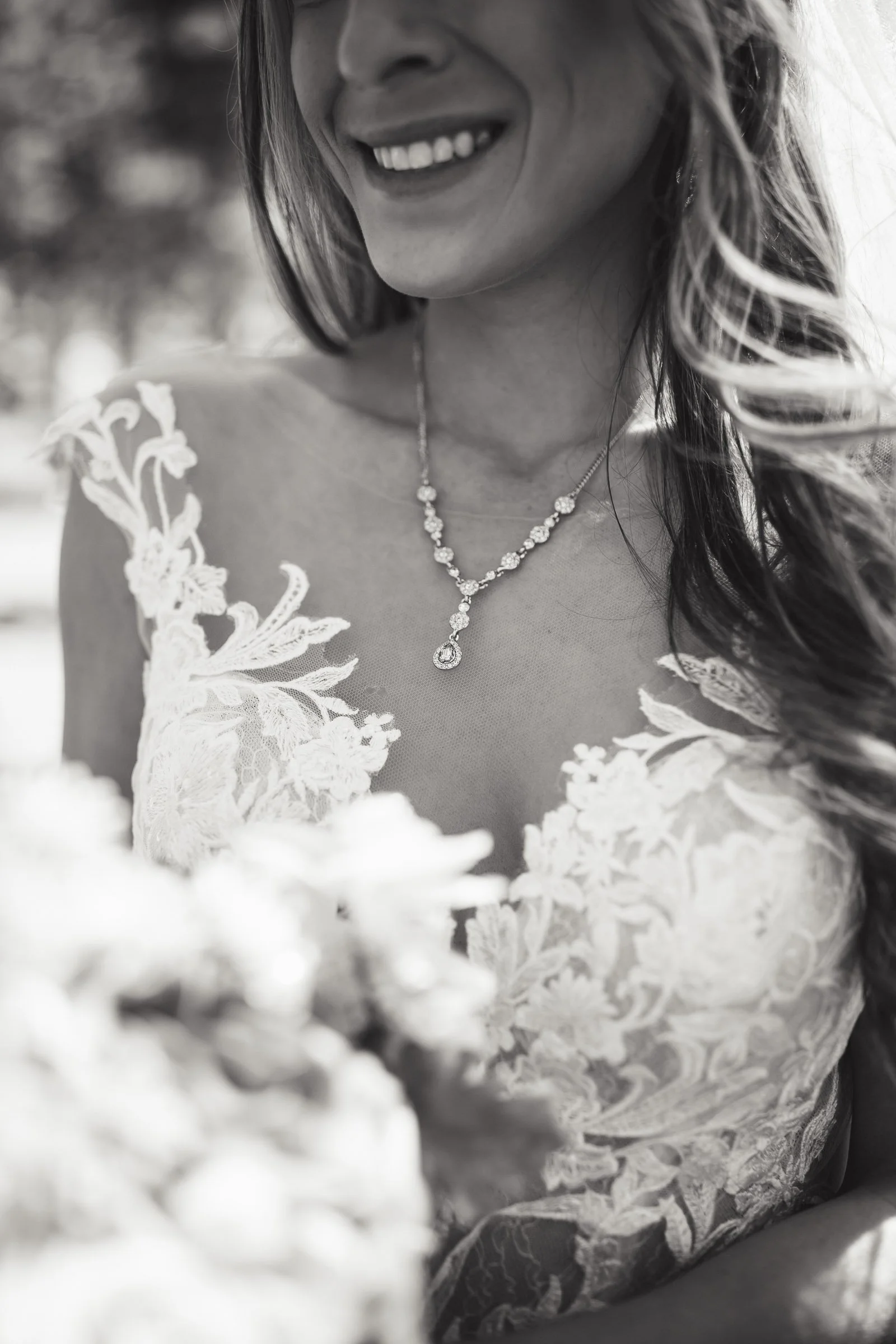 Close-up of a smiling woman in a lace wedding dress wearing a necklace, with an out-of-focus bouquet in the foreground.