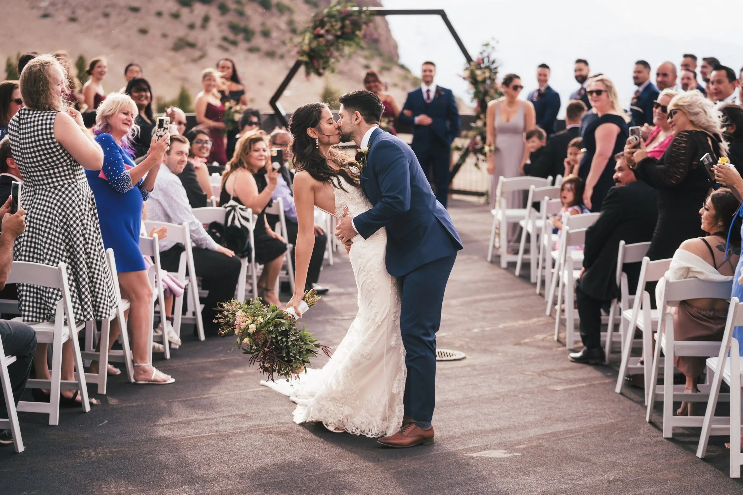A newly married couple kisses during their outdoor wedding ceremony, surrounded by seated guests, with a mountainous background and floral decorations.