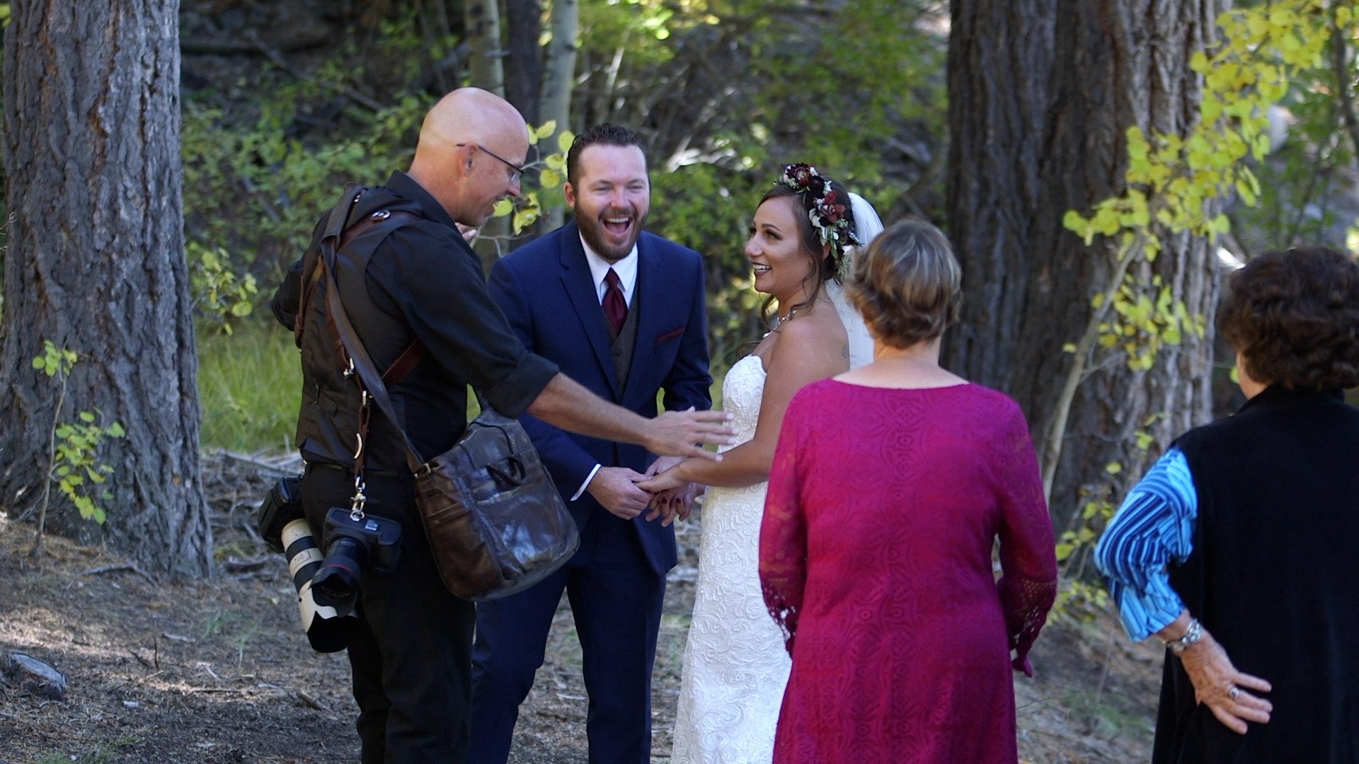 A wedding ceremony taking place outdoors in a wooded area with five people present, including a bride and groom exchanging vows, a photographer taking pictures, and two women watching, one in a magenta dress and one in a black vest with a striped shirt.