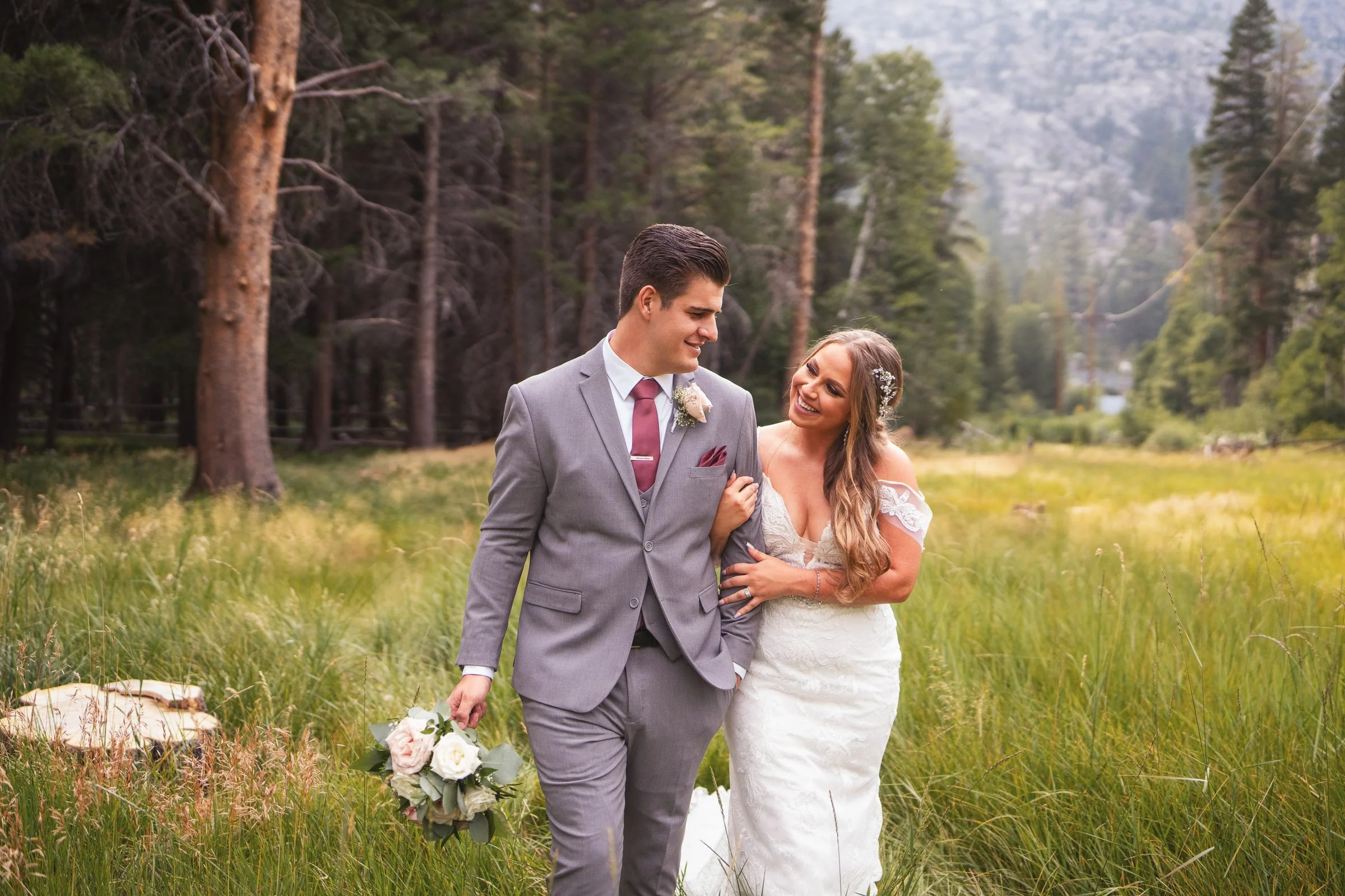 A newlywed couple walking through a grassy field surrounded by tall trees, smiling and looking at each other, with the groom holding a bouquet of flowers.