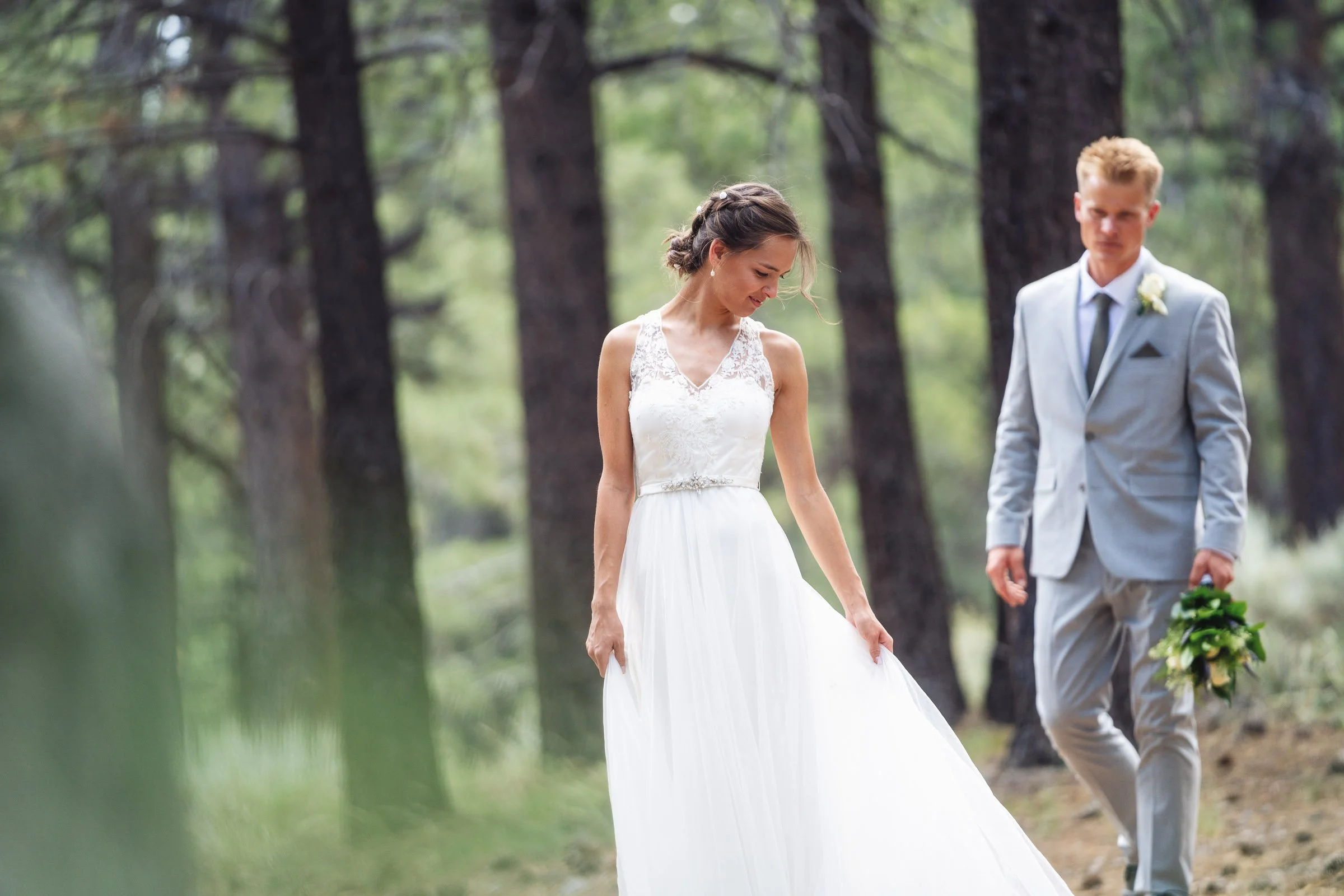 Bride in a white wedding dress looking down with a groom in a light grey suit holding a bouquet of flowers walking behind her in a forest setting.