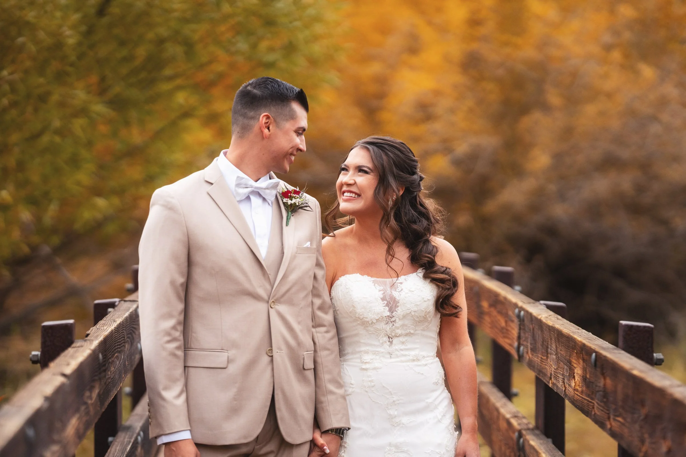 A bride and groom stand on a wooden bridge outdoors during fall, holding hands and smiling at each other, with autumn-colored trees in the background.