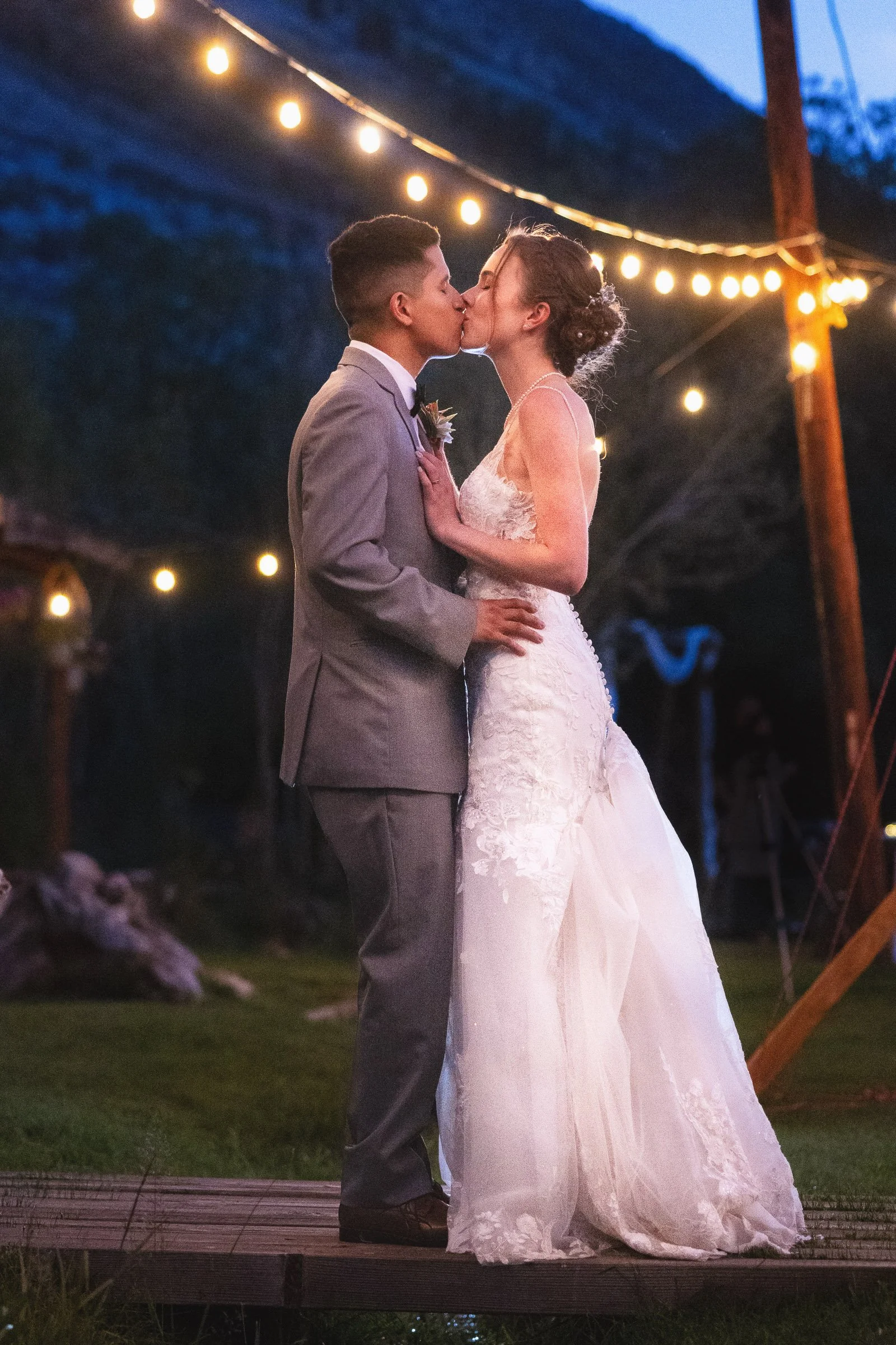 A newlywed couple shares a kiss outdoors at night, under string lights, with a mountain backdrop.
