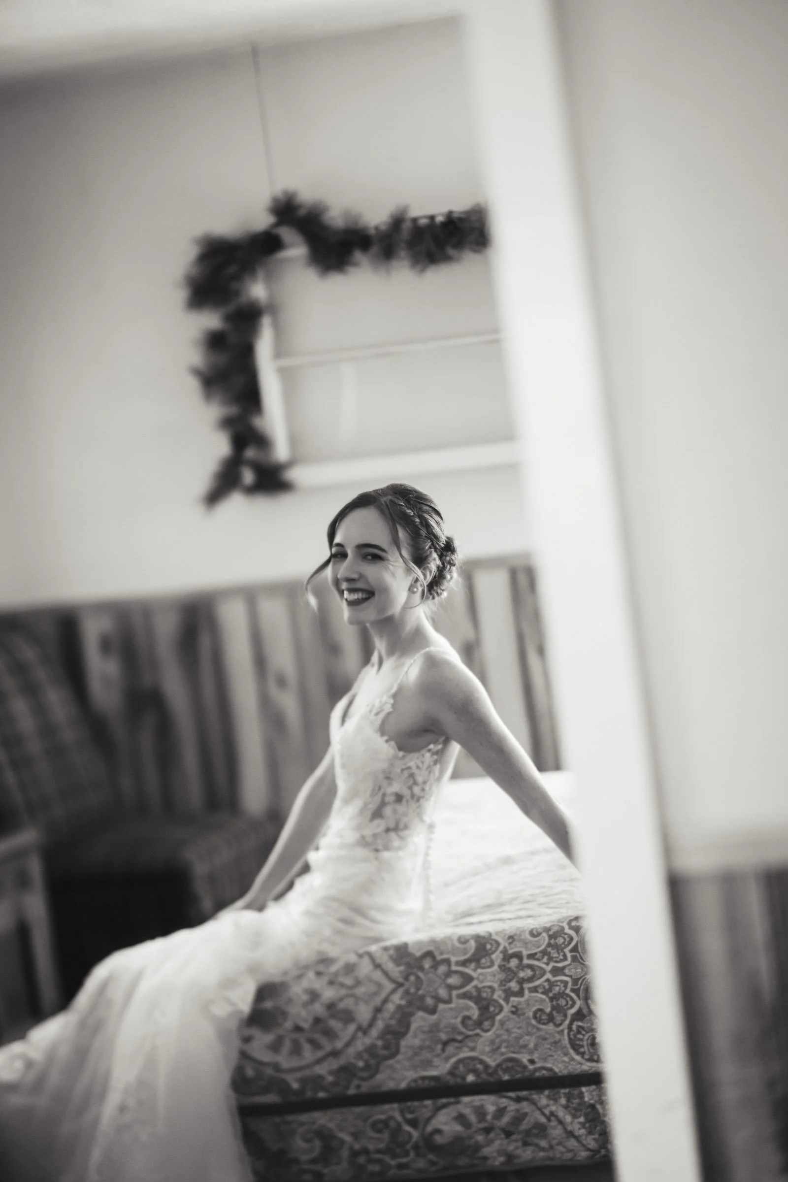 A smiling woman in a wedding dress sitting on a cushioned bench, reflected in a mirror. The photo is in black and white.