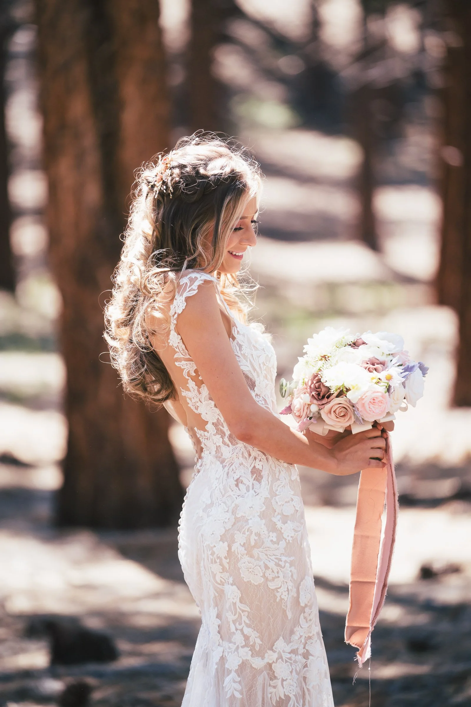 A bride in a lace wedding dress holding a bouquet of white and pink flowers, standing outdoors in a wooded area, smiling and looking down.