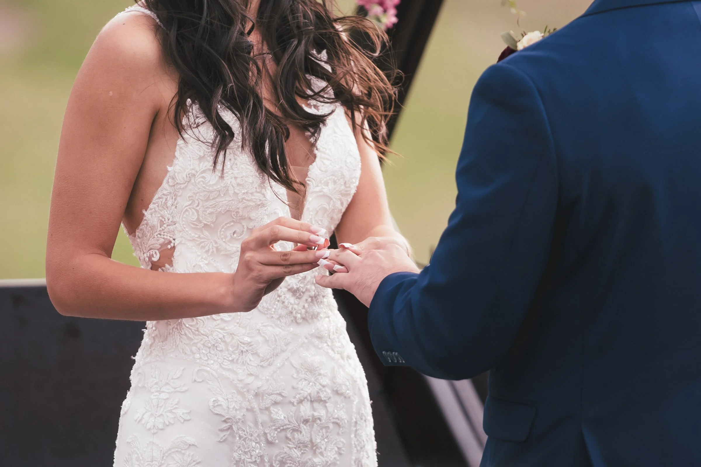 A bride wearing a white lace wedding dress during a wedding ceremony, placing a ring on the groom's finger, who is dressed in a navy blue suit.
