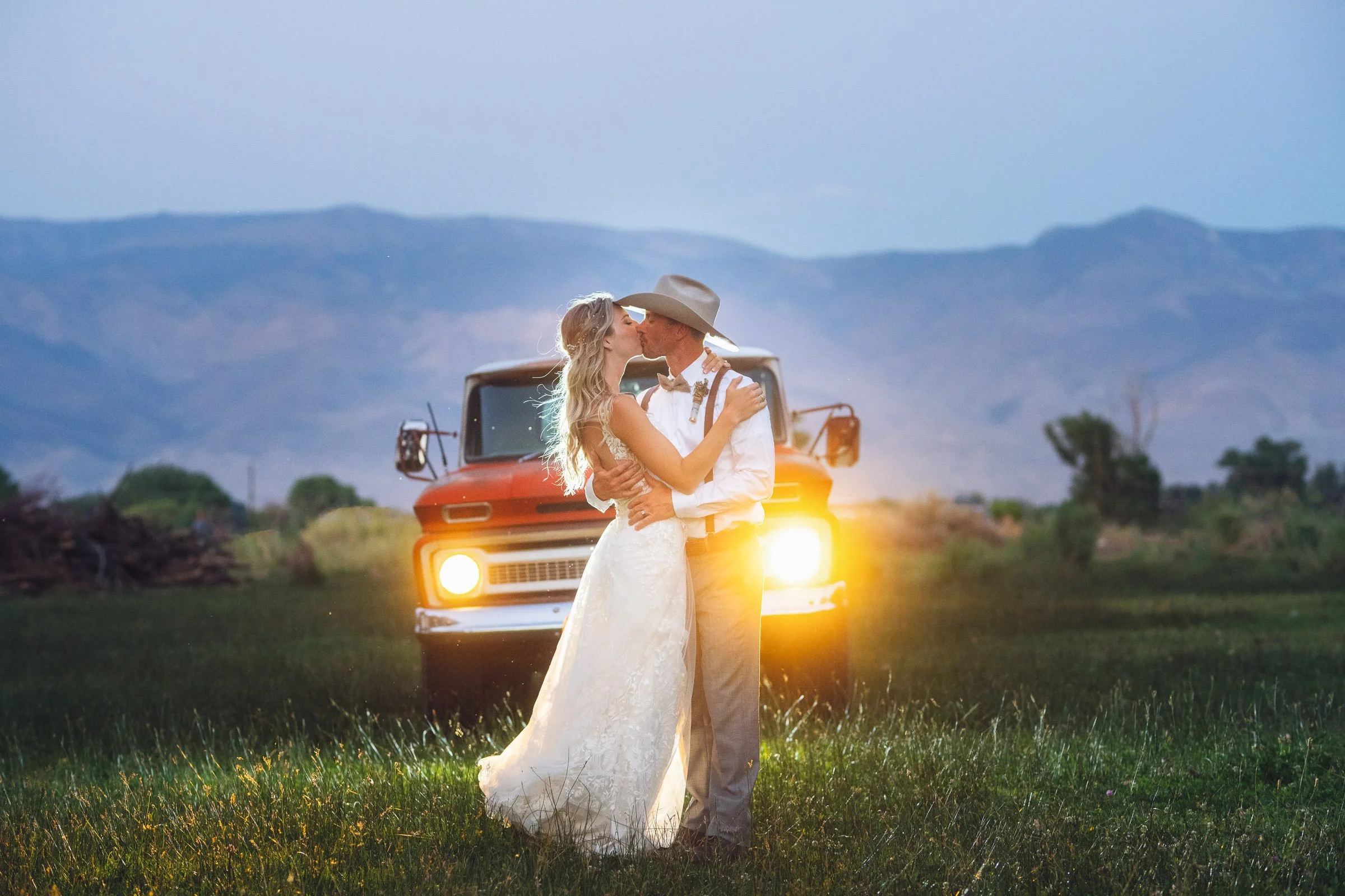 A newlywed couple sharing a kiss in a scenic outdoor landscape during sunset or twilight, with a lake, mountains, and trees in the background.