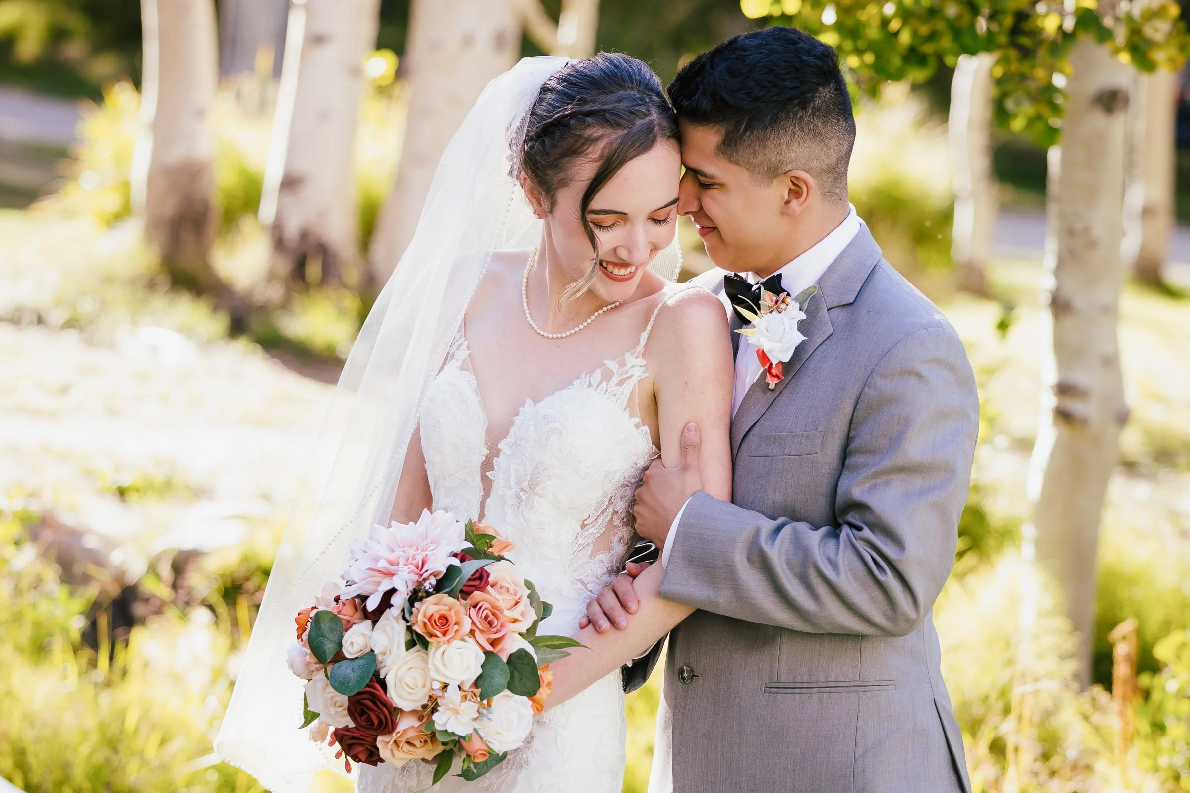 A bride and groom embracing outdoors, smiling with foreheads touching, the bride holding a bouquet of roses and peonies, surrounded by trees and sunlight.
