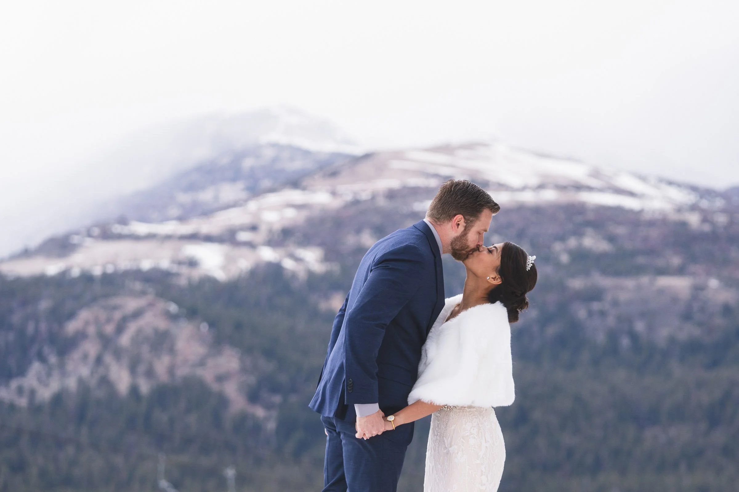 A newlywed couple sharing a kiss outdoors with snowy mountains in the background.