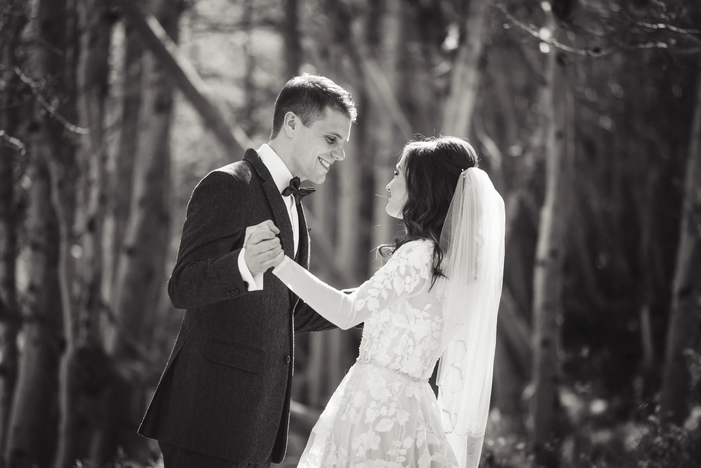 A black and white photograph of a bride and groom dancing together outdoors, with trees in the background. The groom is wearing a suit with a bowtie, and the bride is dressed in a lace wedding gown with a veil.