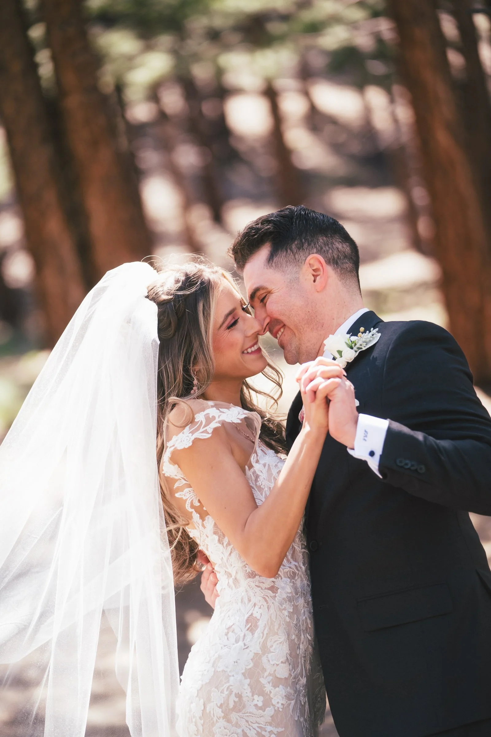 A bride and groom smiling and holding hands in a forest with tall trees, close together with their noses touching.