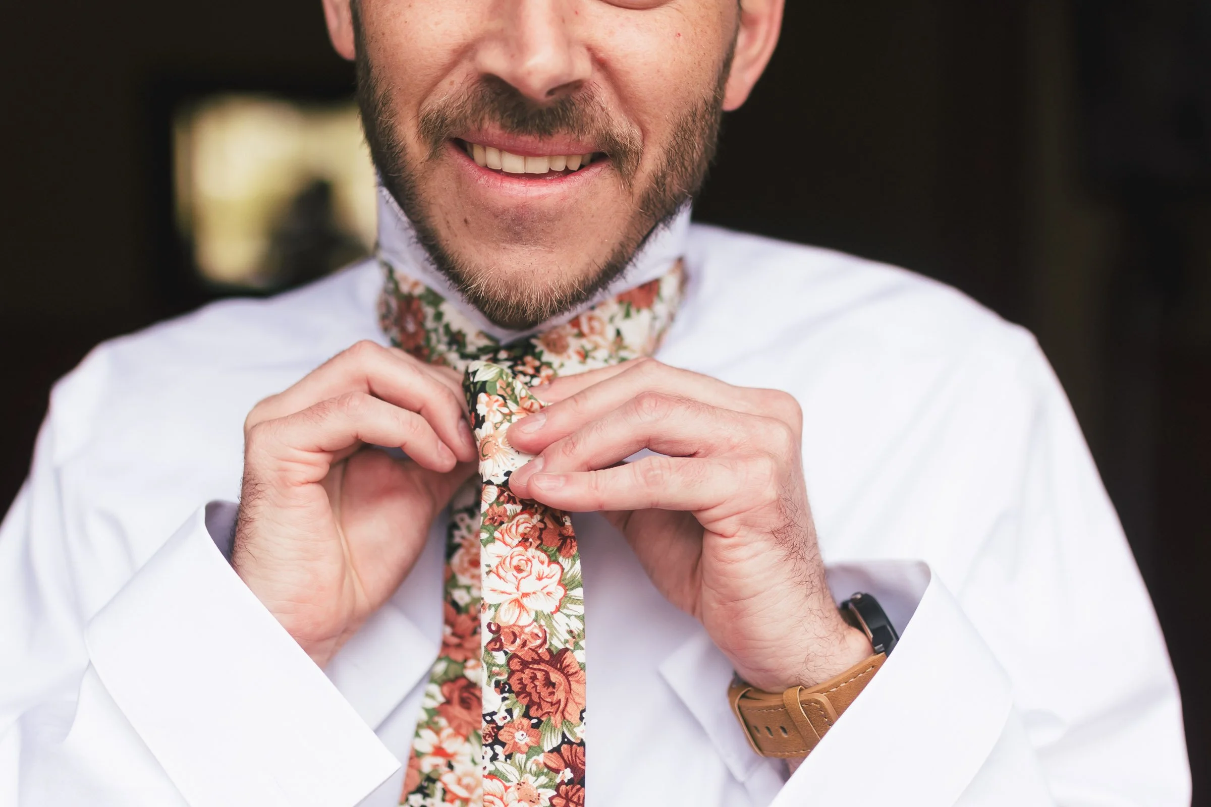 A man smiling as he ties a floral patterned tie, wearing a white shirt and a wristwatch.